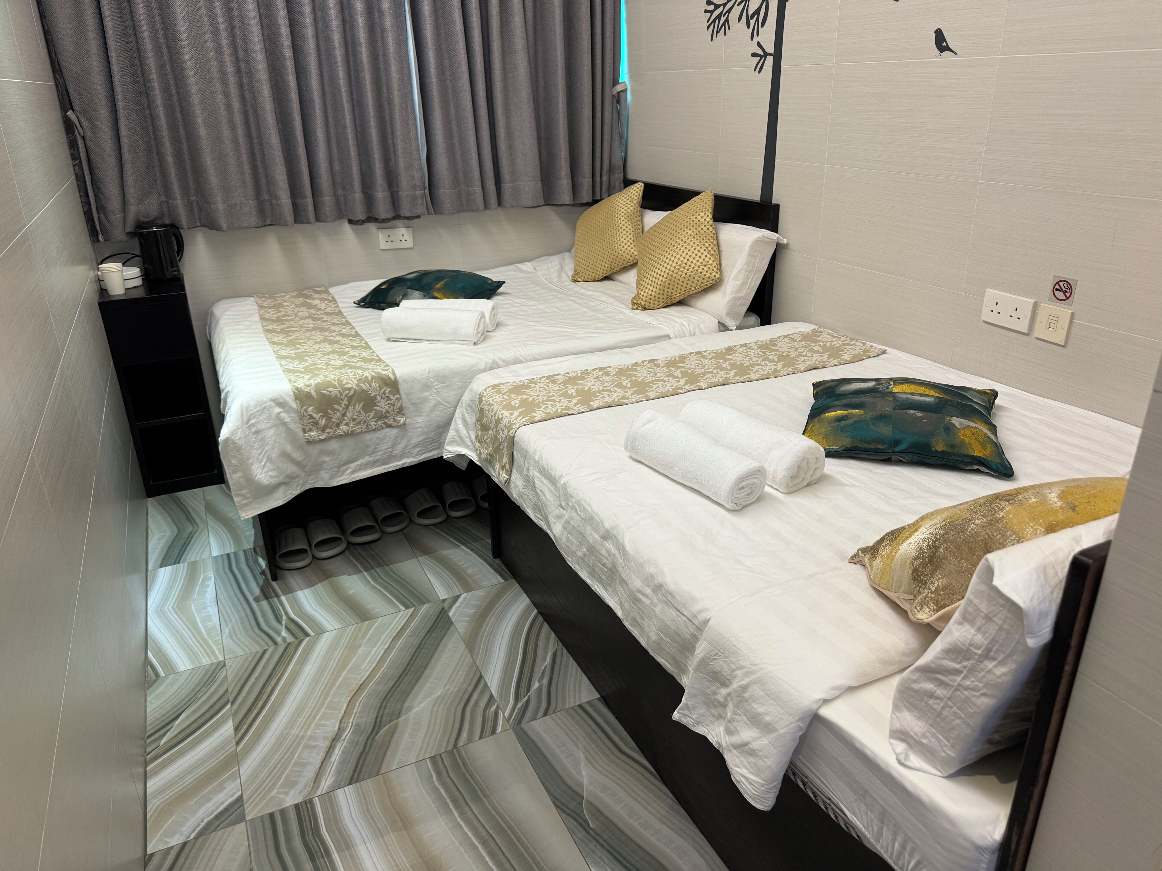 A hotel room with two beds, grey curtains, a coffee maker, and slippers on the floor.