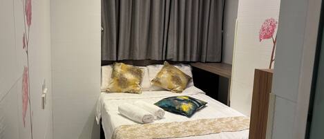 Un dormitorio pequeño y ordenado con cama, almohadas, cabecera de madera y cortina.
