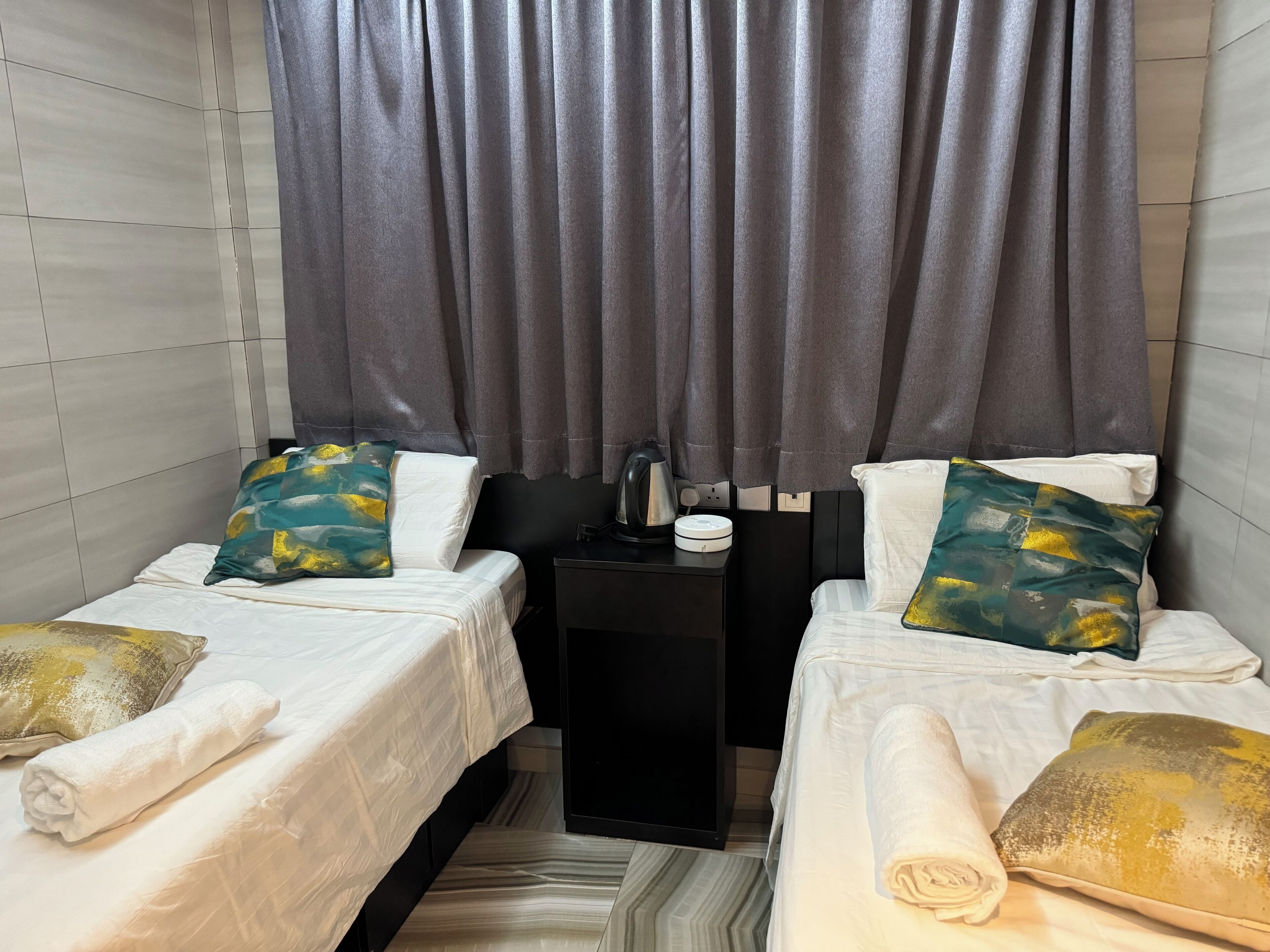 A small, modern room with two beds, grey curtains, a black nightstand with a kettle, and a patterned rug.