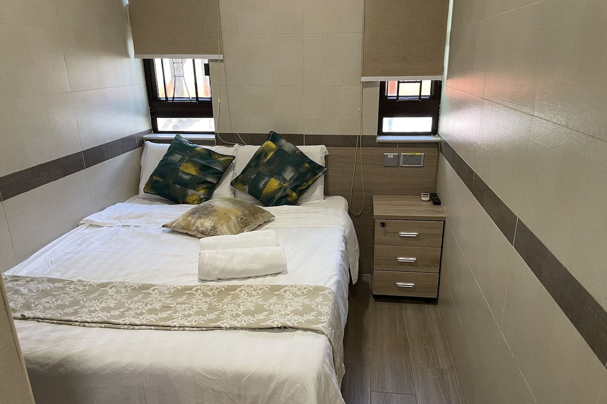 A compact hotel room with a bed, two bedside tables, a small drawer unit, and a window with blinds.