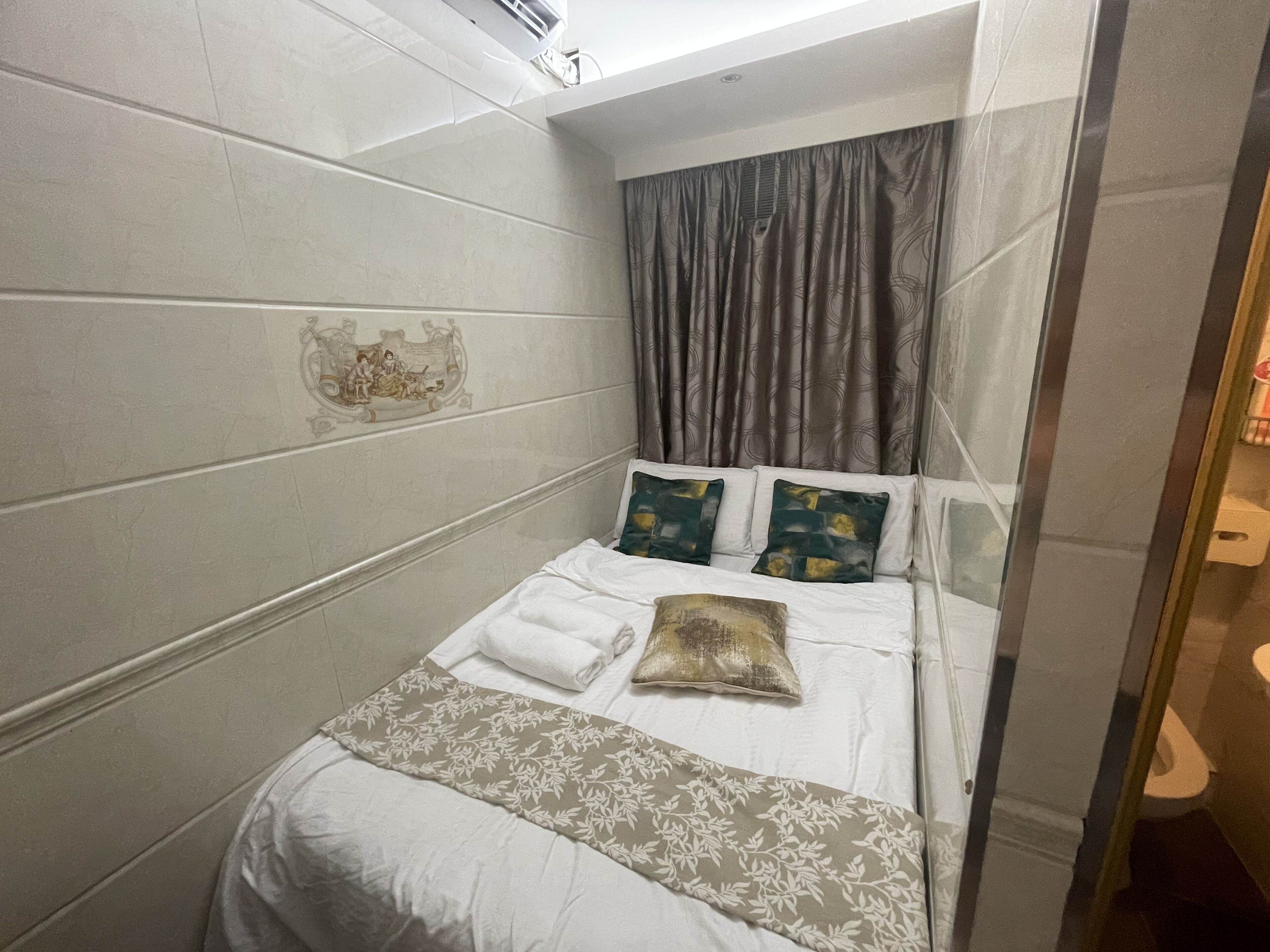 A small, neatly made bed with white bedding and a floral-patterned throw, a single decorative pillow, and a wall-mounted air conditioning unit.