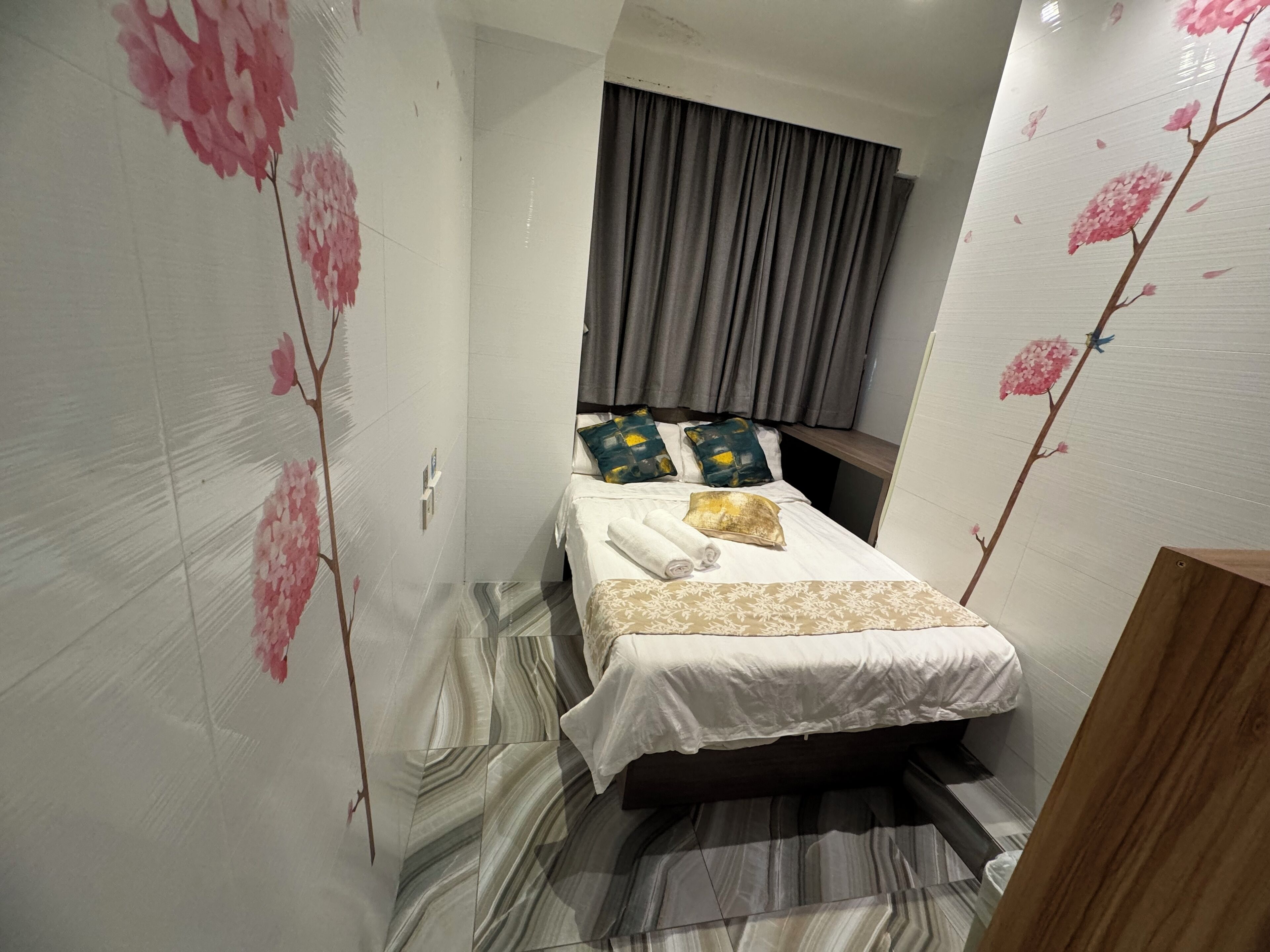 A narrow hotel room with a bed, a wooden headboard, and a curtain.