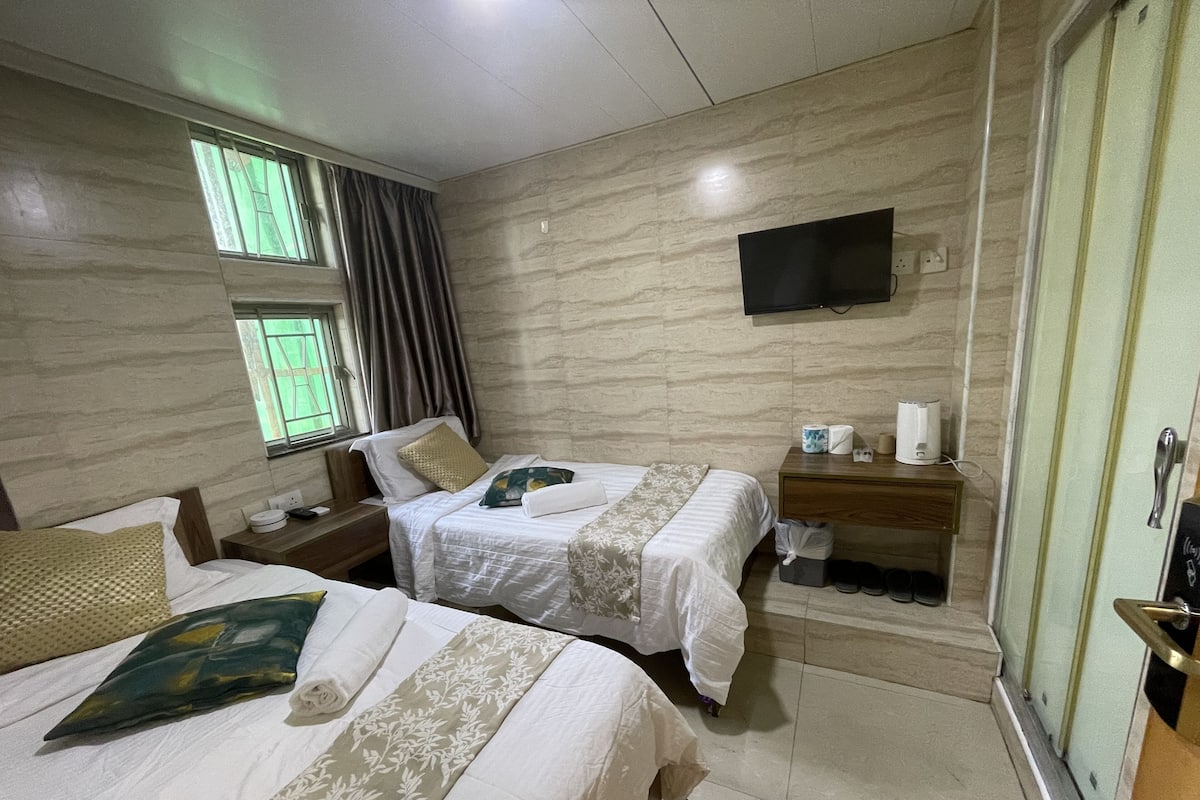 A hotel room with two beds, a TV mounted on the wall, a bedside table with a lamp, and a window with curtains.