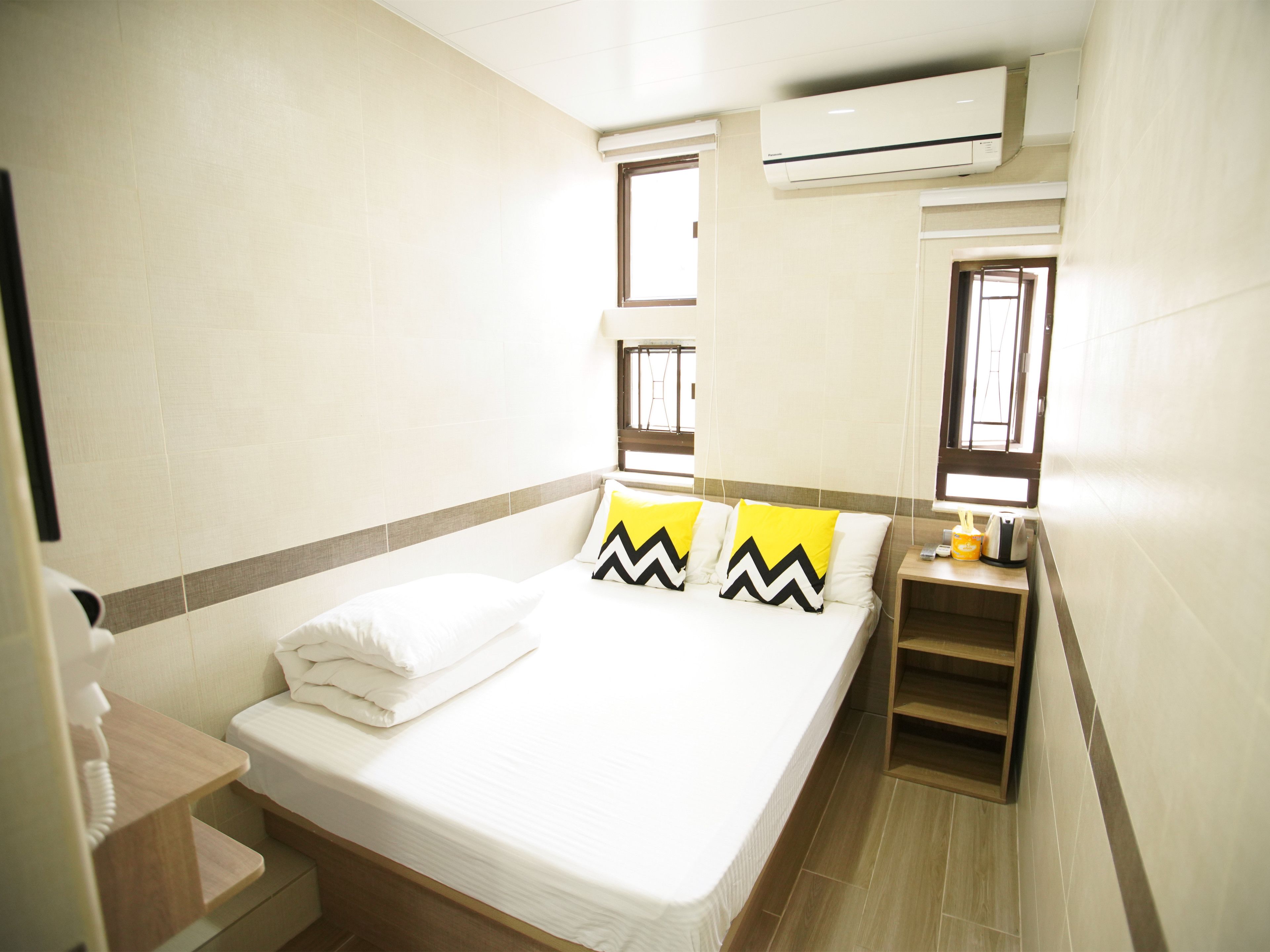 Une chambre d’hôtel avec un lit, deux fenêtres, un appareil de climatisation, une table de chevet et une petite étagère.