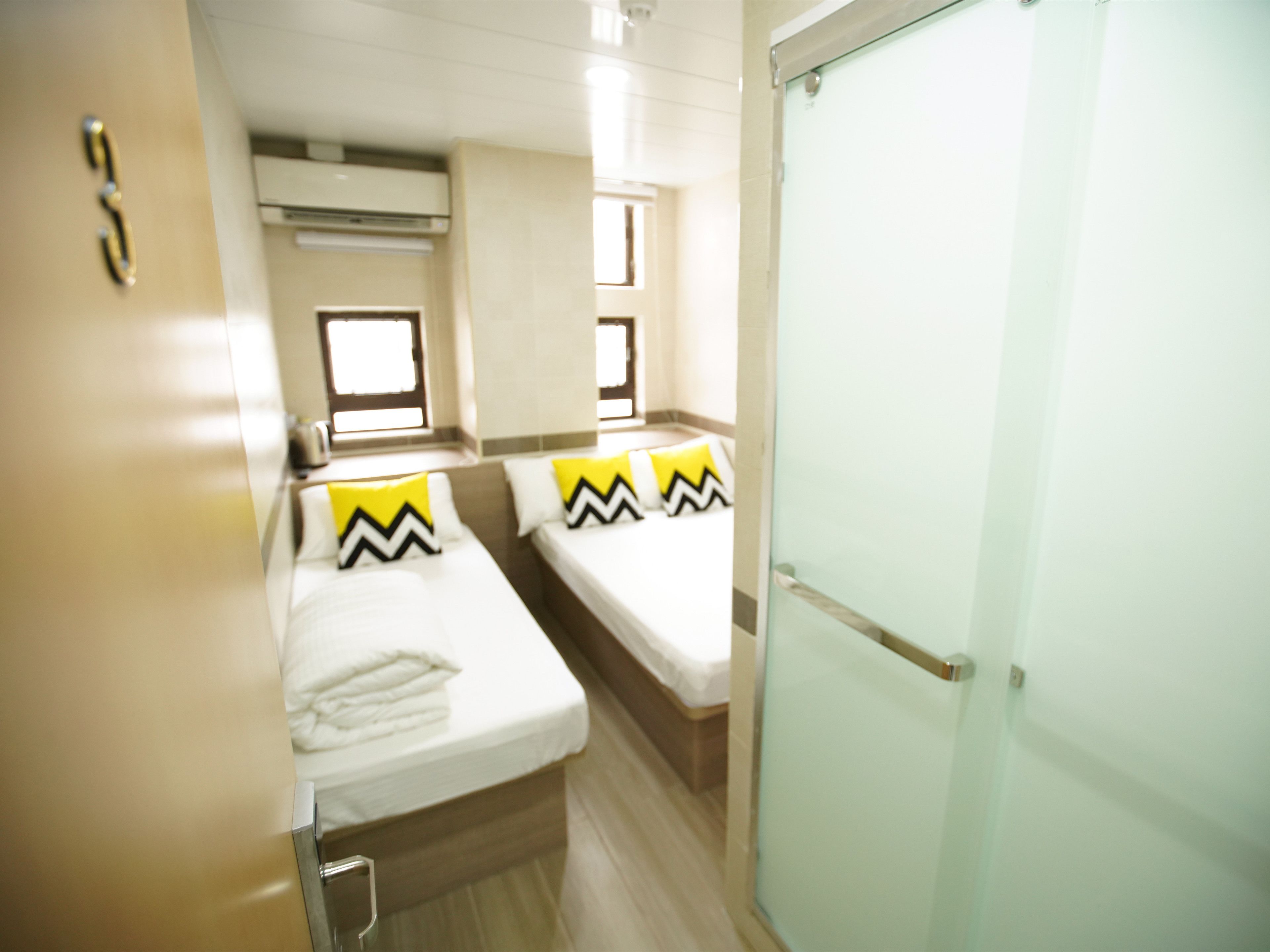 Ein Hotelzimmer mit zwei Betten, jedes mit einer weißen Matratze und einem gelb-schwarz gestreiften Kissen. Es gibt ein Fenster mit Vorhängen, eine Klimaanlage über den Betten sowie eine Tür mit Glasfenster.