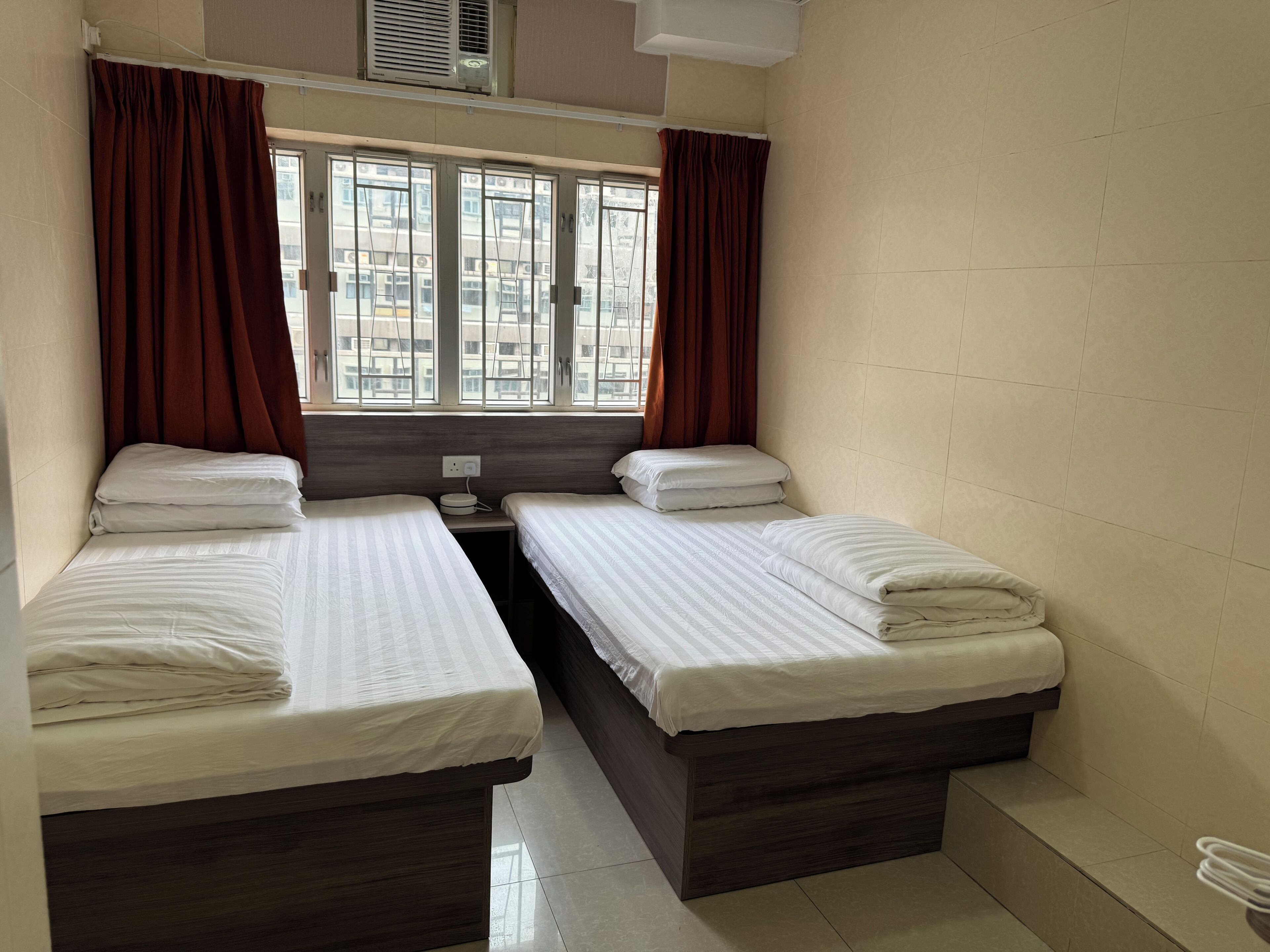 A small, clean room with two single beds, a nightstand, and a window with curtains.