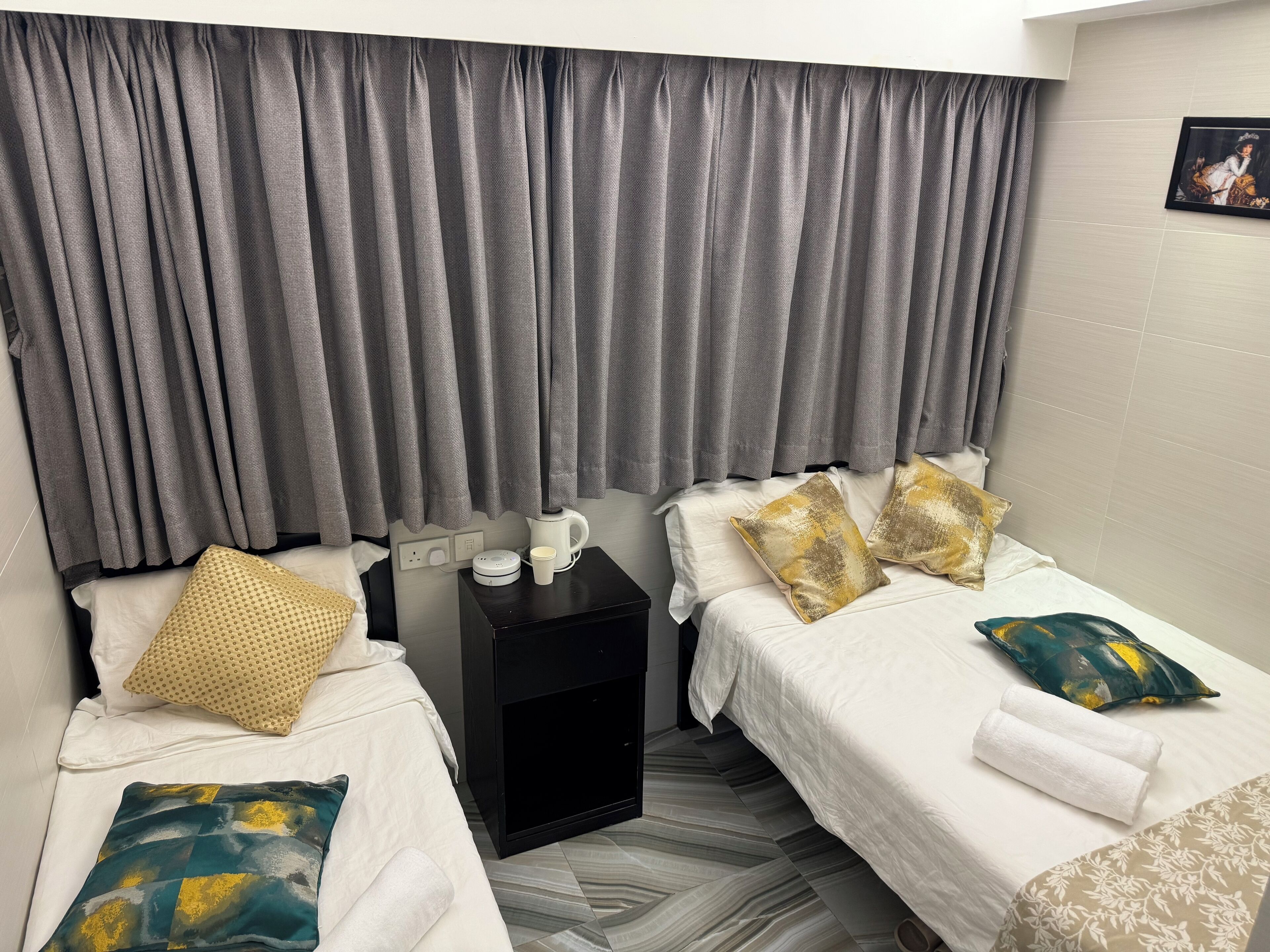 A hotel room with two beds, grey curtains, a black nightstand with a kettle and a cup, and patterned pillows.