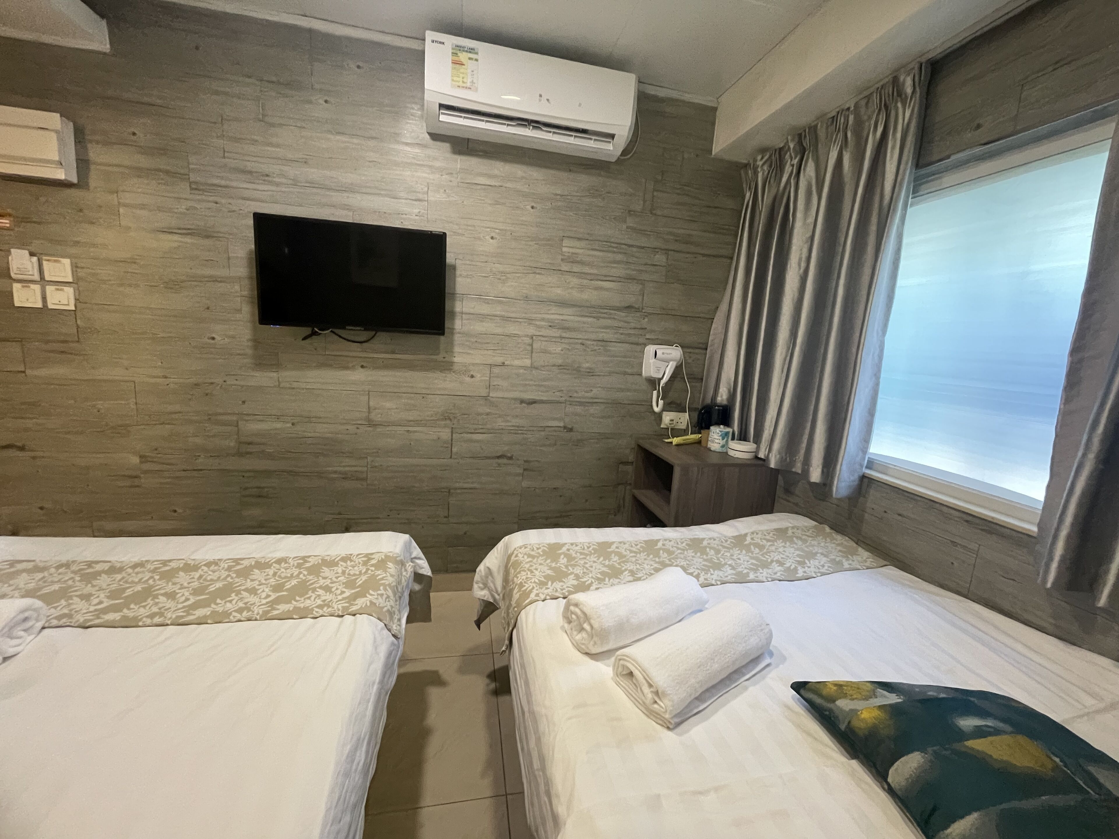 Ein Hotelzimmer mit zwei Betten, einem flachbildfernseher an der Wand, einer Klimaanlage, einem Nachttisch mit Telefon und Lampe sowie einem Fenster mit Vorhängen.
