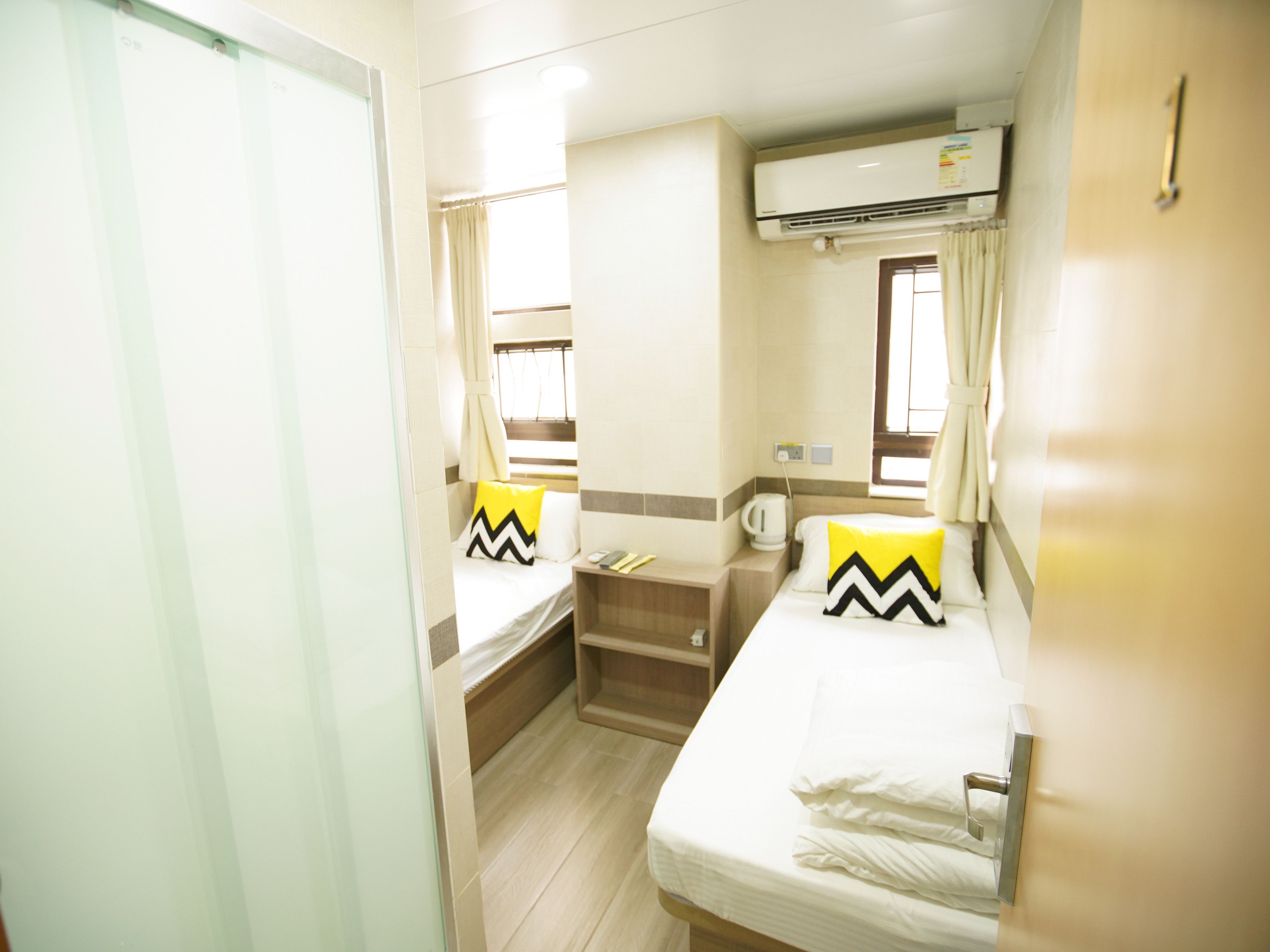 A compact hotel room with two single beds, a small seating area, a window with curtains, an air conditioning unit, and a kitchenette.