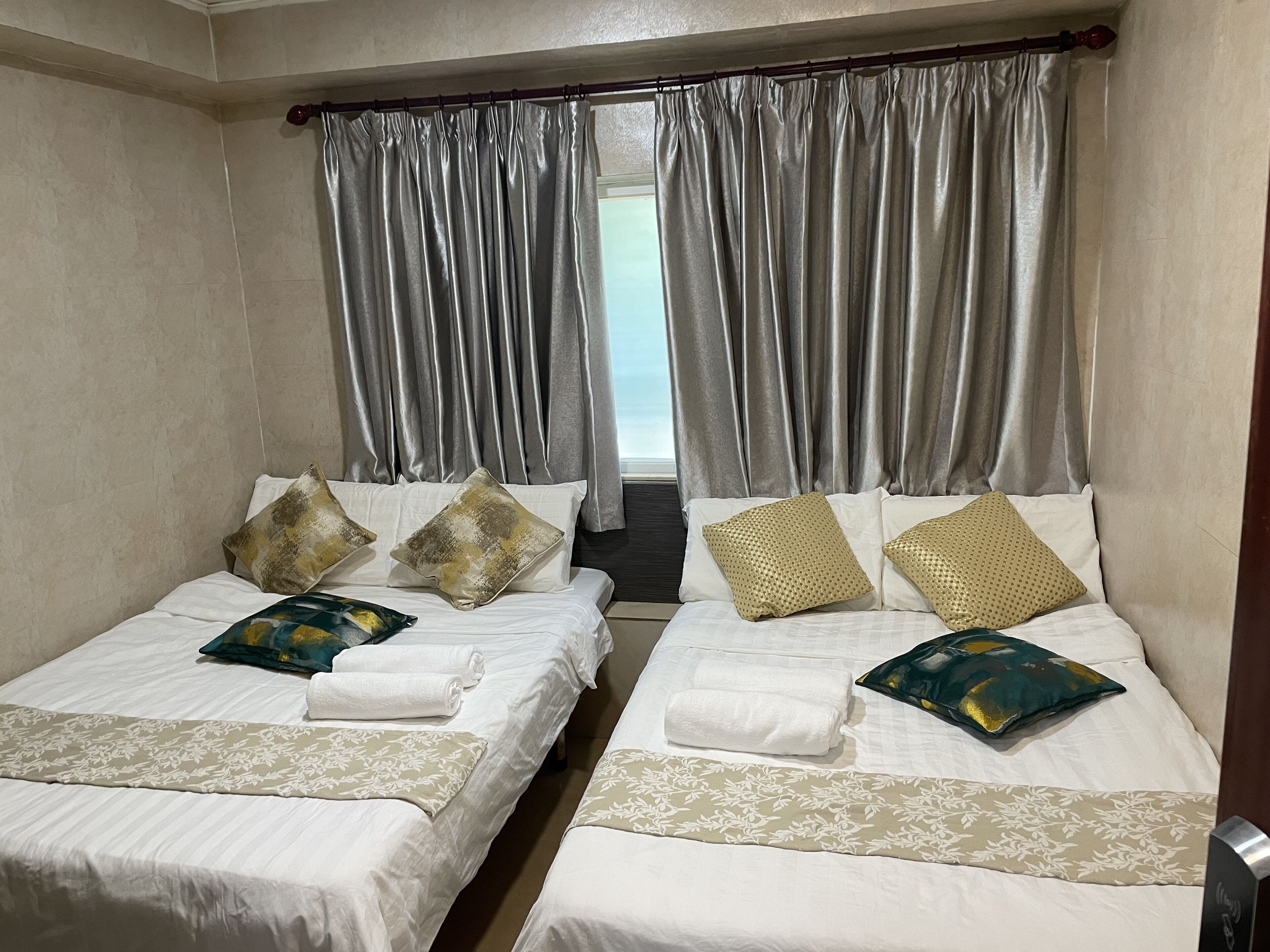 A hotel room with two beds, each with white linens and decorative pillows. The room has a large window with grey curtains, and a view of the outside.