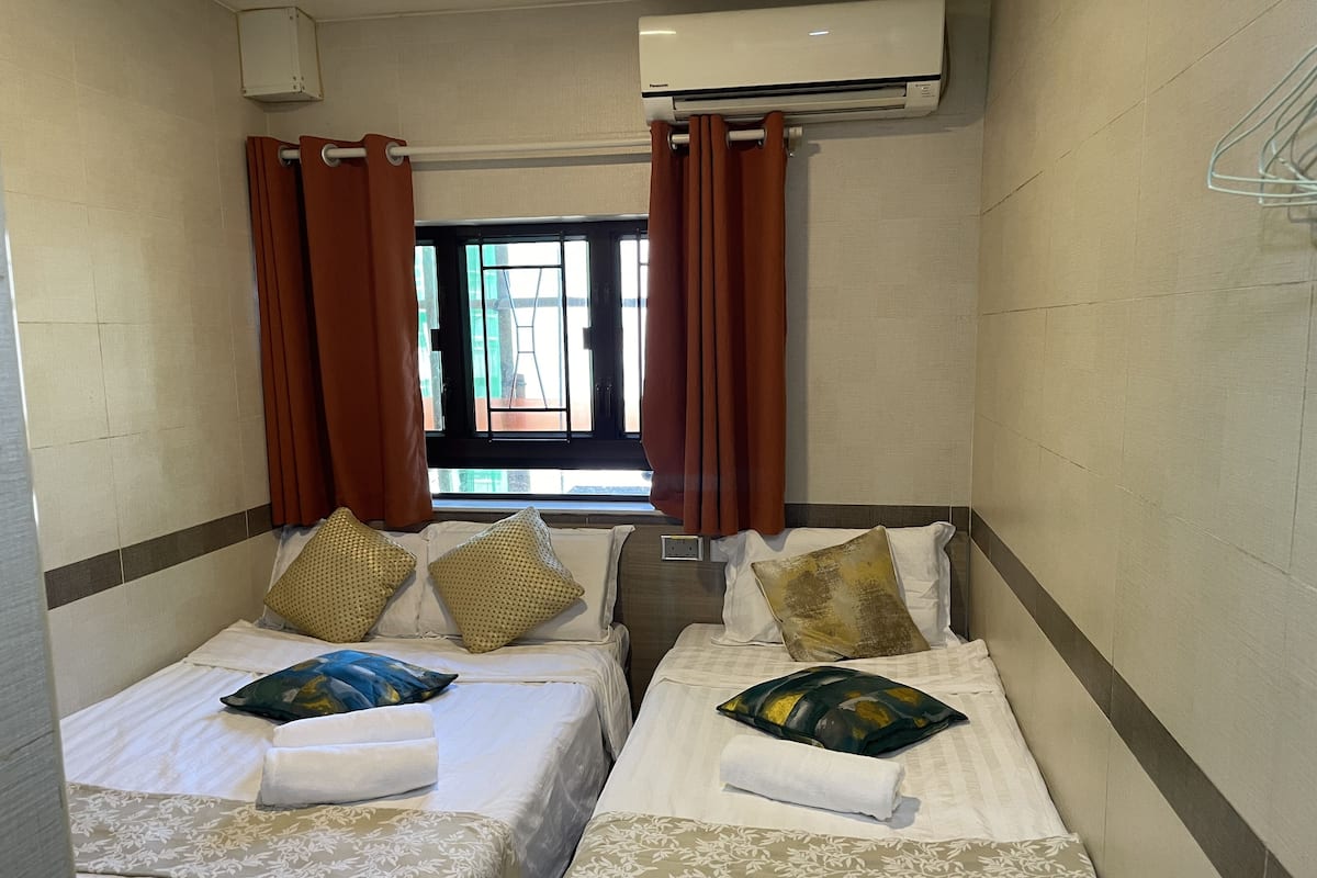 A hotel room with two beds, brown curtains, a window, and an air conditioning unit.