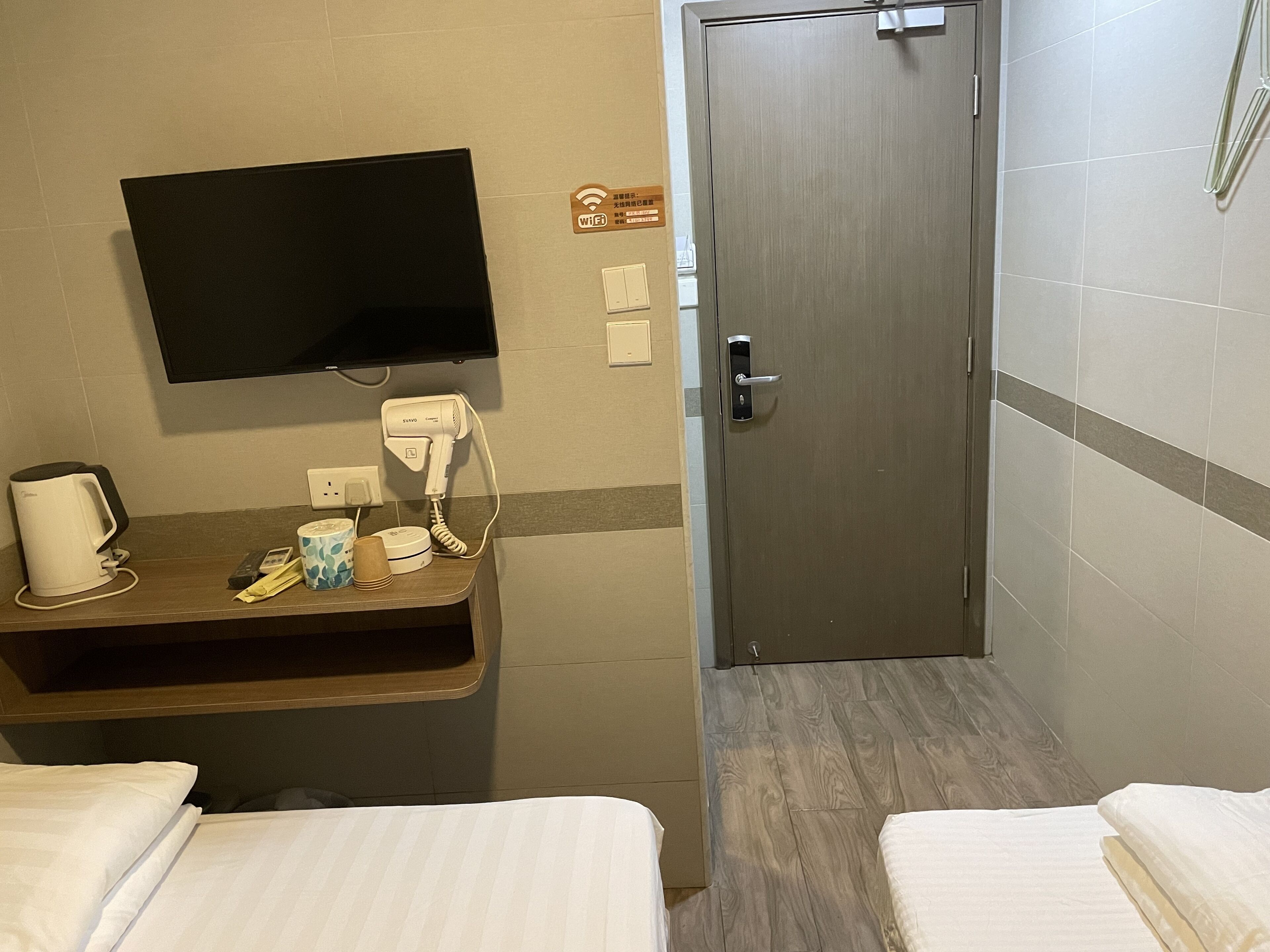 A hotel room with a flat-screen TV mounted on the wall, a hairdryer, a coffee maker, and a small table with toiletries. There are two beds with white linens and a wooden floor.