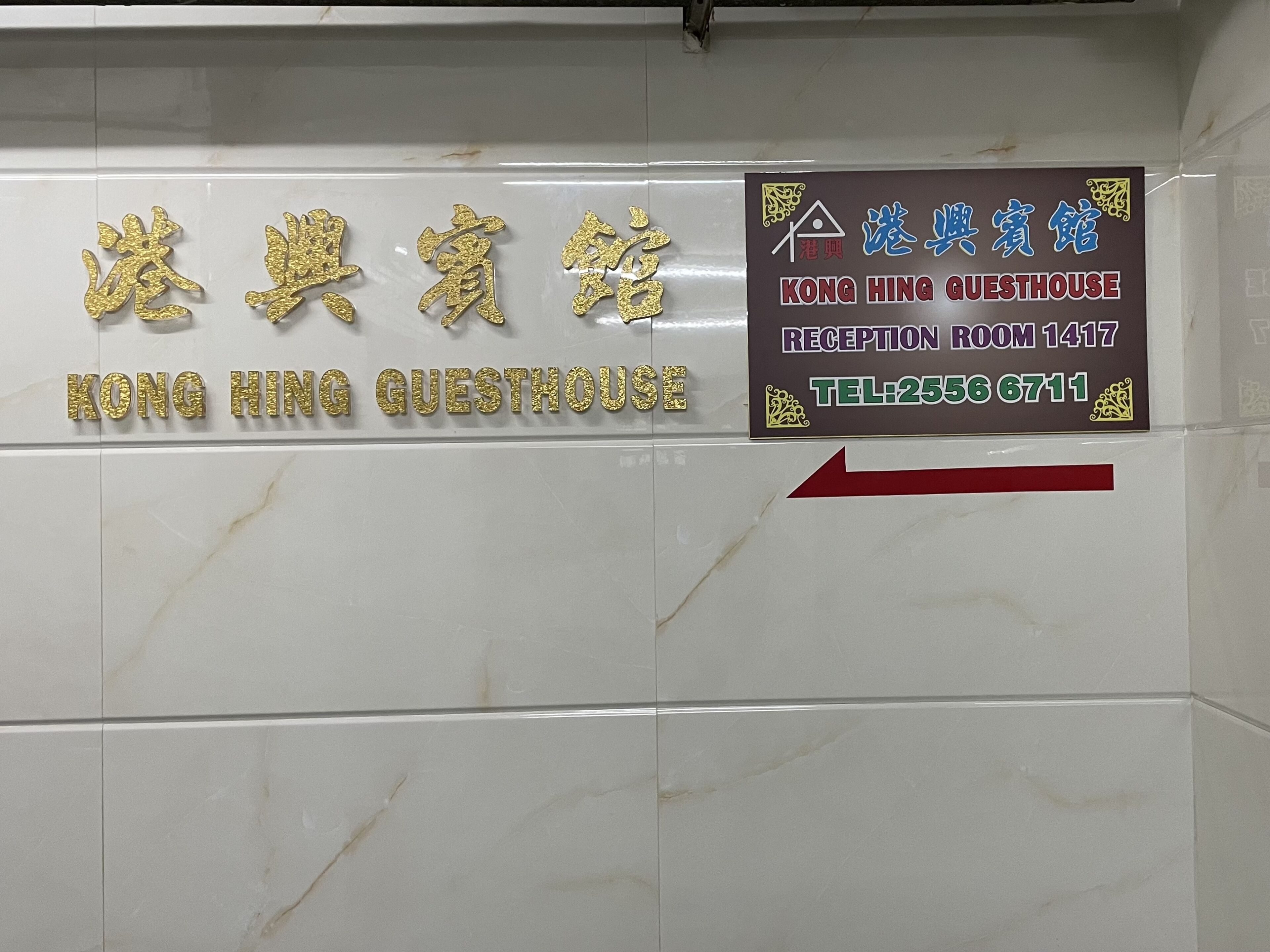 A hotel sign with the name 'Kong Hing Guesthouse' in gold letters on a white wall. There is a red arrow pointing to the right.