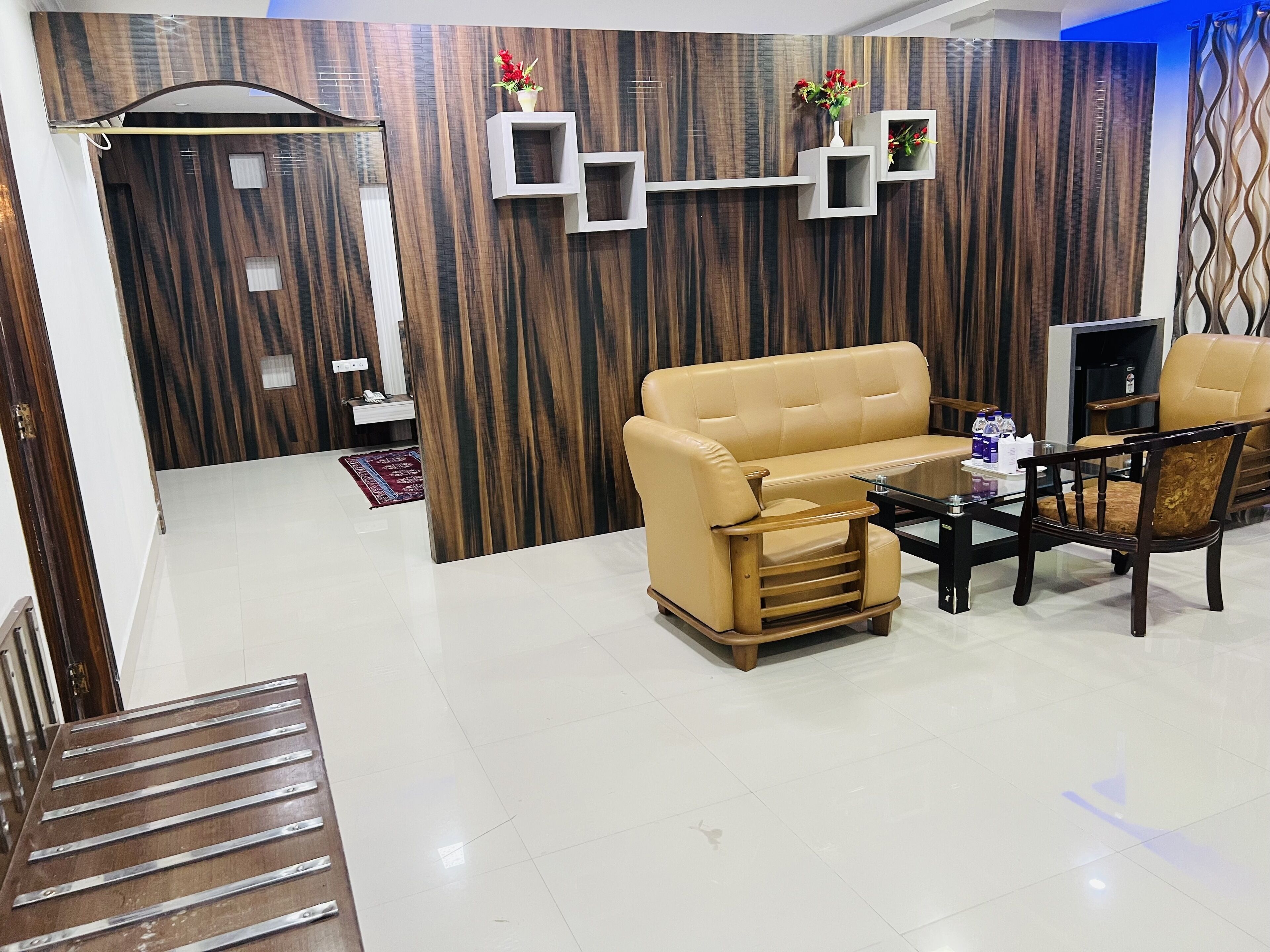A modern living room with a beige sofa, a glass coffee table, and wooden chairs. There is a built-in shelf with decorative items and a wooden accent wall.