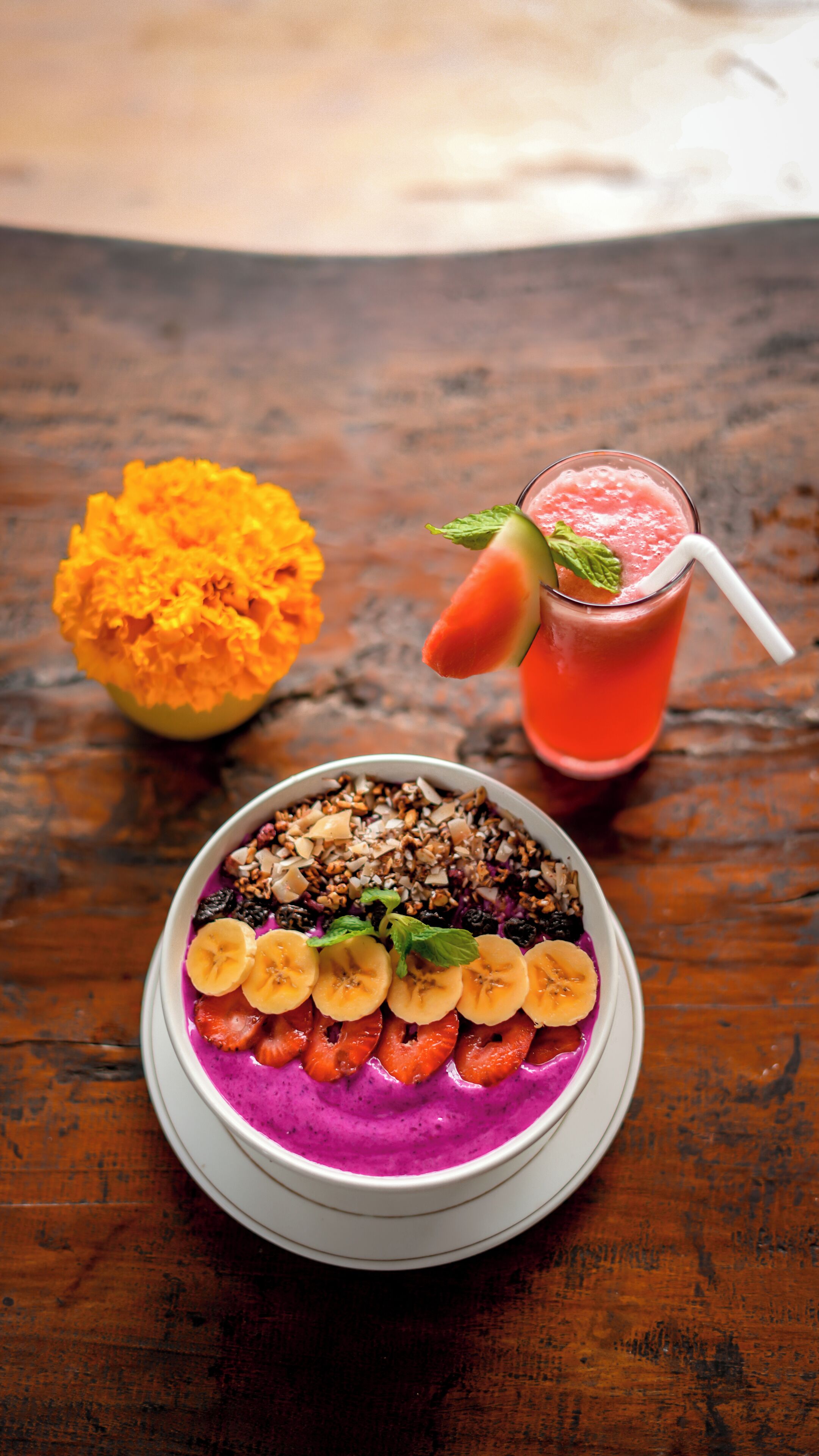 A bowl of smoothie topped with banana slices and strawberries, a glass of smoothie with a slice of watermelon and mint, and a bowl of granola with nuts and dried fruits on a wooden table.