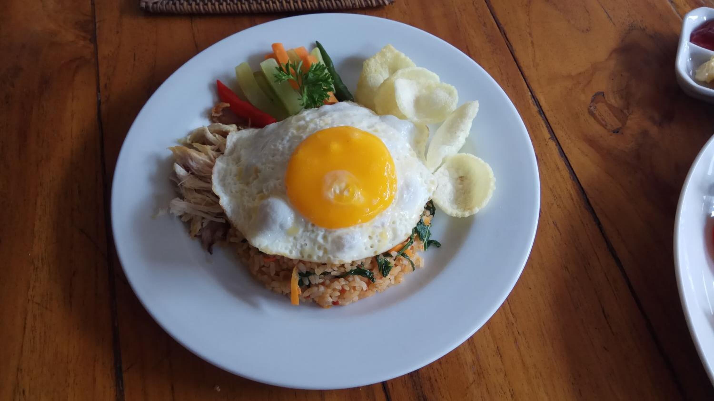 A plate of food with a fried egg on top, accompanied by sliced bananas, a side of vegetables, and a small bowl of sauce.