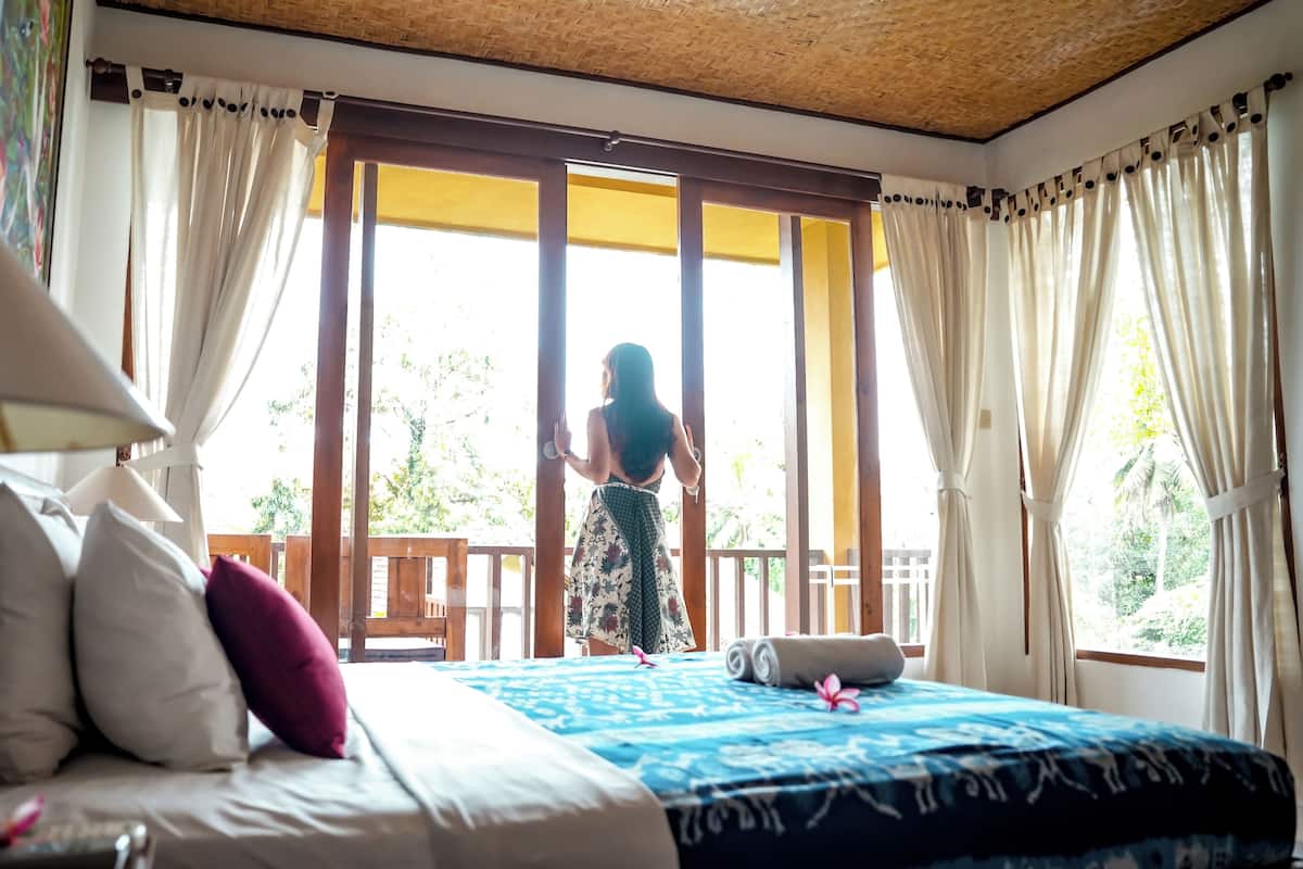 A room with a bed, a nightstand, a lamp, and a view of a balcony with a person standing outside.