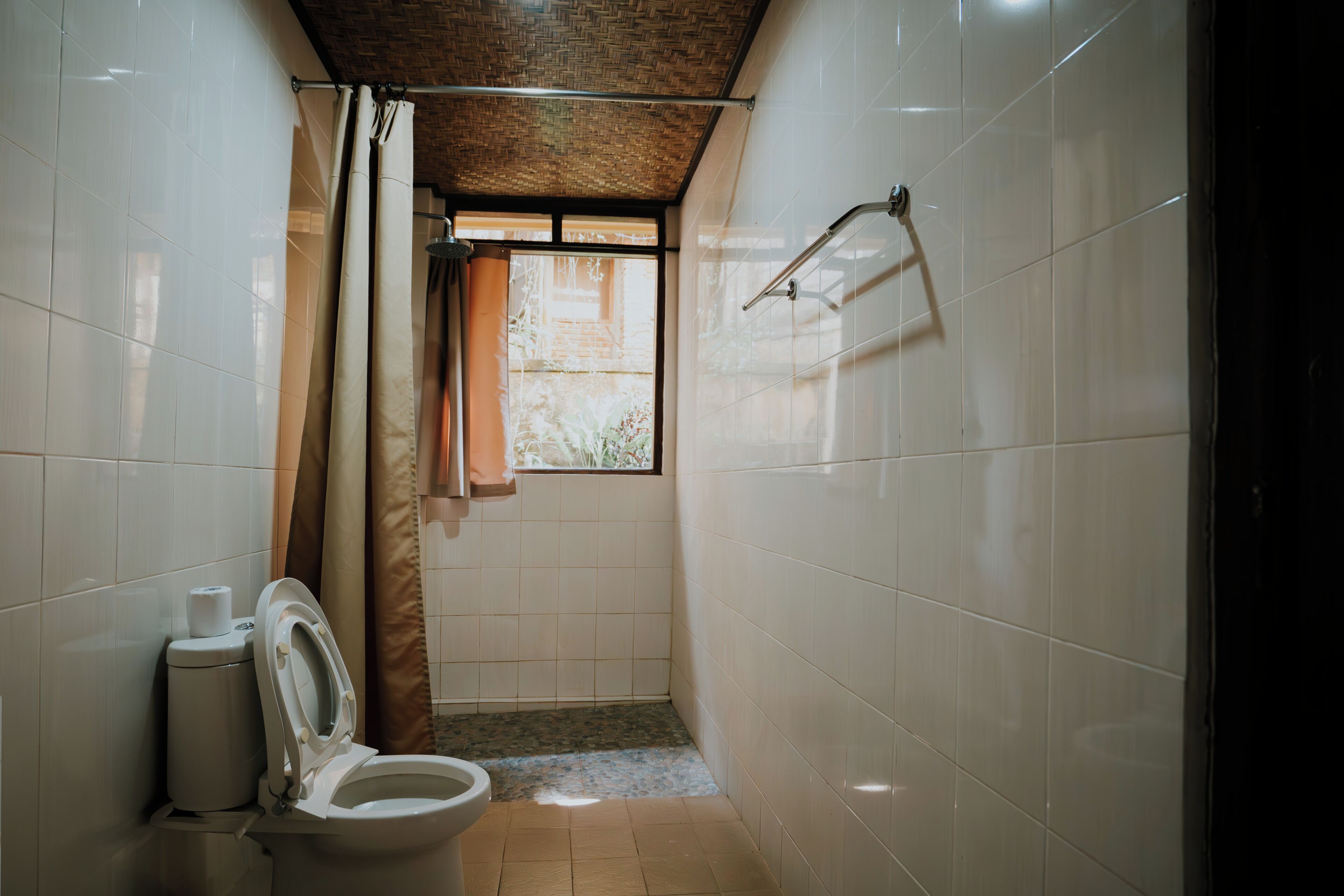 Un baño pequeño y estrecho con azulejos blancos, un inodoro, una cortina de ducha y una ventana con vistas al verdor.