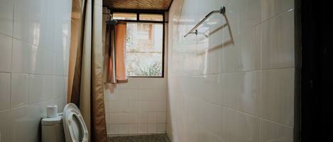 A small, narrow bathroom with white tiles, a toilet, a shower curtain, and a window with a view of greenery.