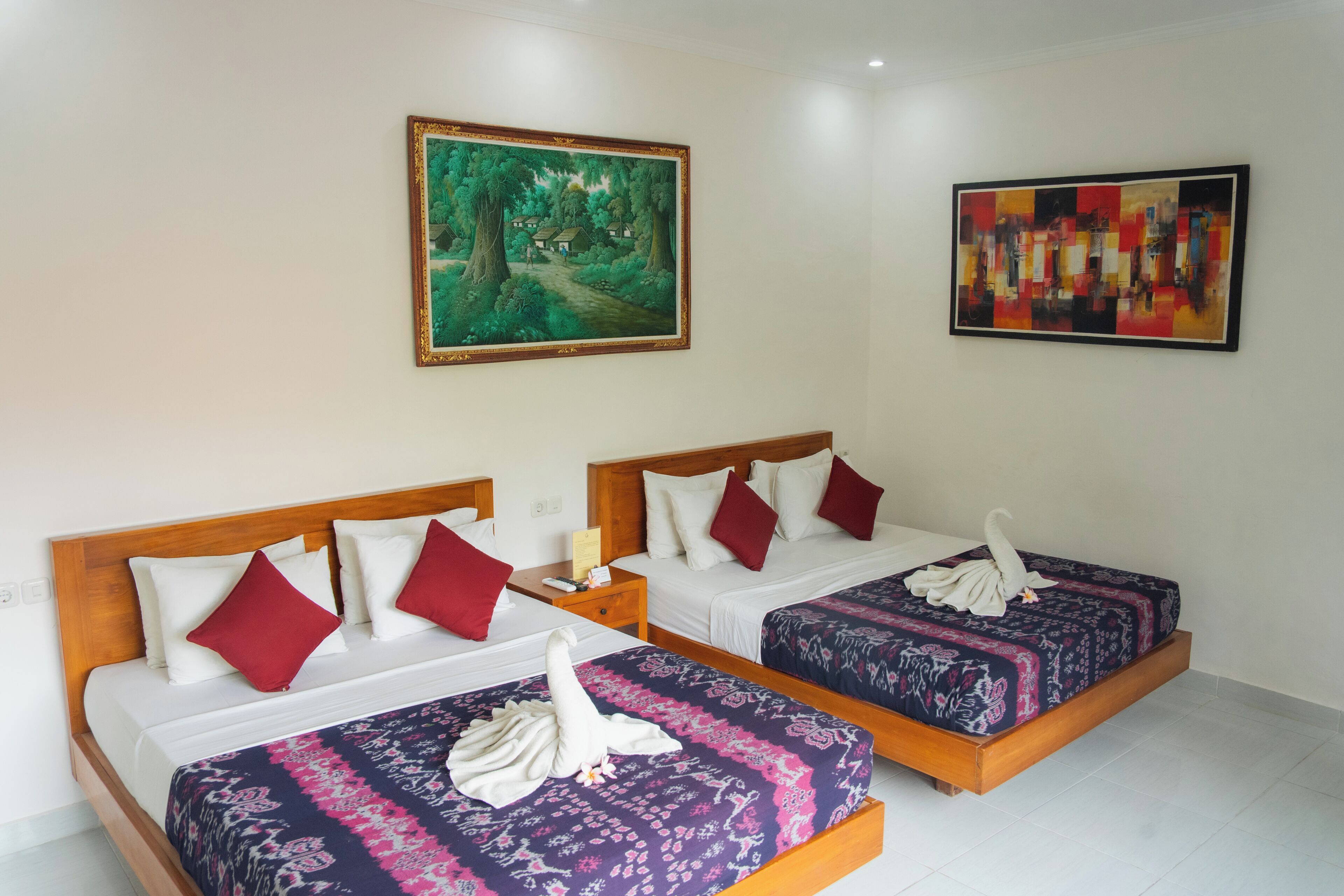 Habitación de hotel con dos camas, cada una con cabecero de madera, ropa de cama blanca y almohadas rojas. Hay dos cuadros en las paredes, uno que representa un paisaje forestal y el otro una composición abstracta.