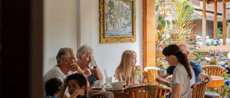 Eine Gruppe von Menschen speist in einem Straßencafé mit Holztischen und -stühlen, einem Fliesenboden und einer verzierten Decke.