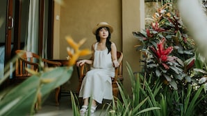 A woman sitting on a chair in a room with a wooden ceiling, surrounded by lush green plants and a large window. - Jati 3 Bungalows (Ubud)