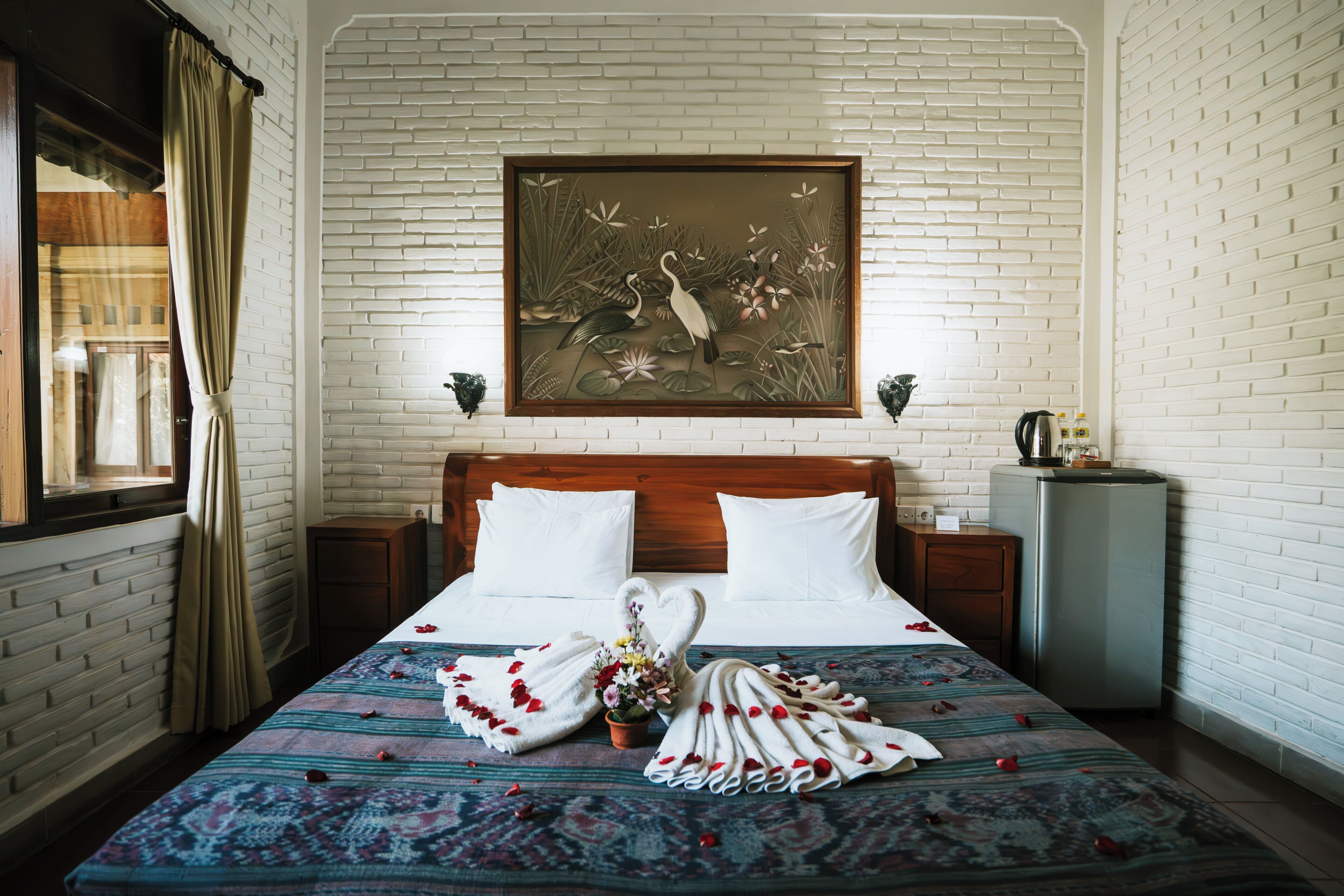 A hotel room with a bed, a nightstand, a wall-mounted painting, and a window with curtains.