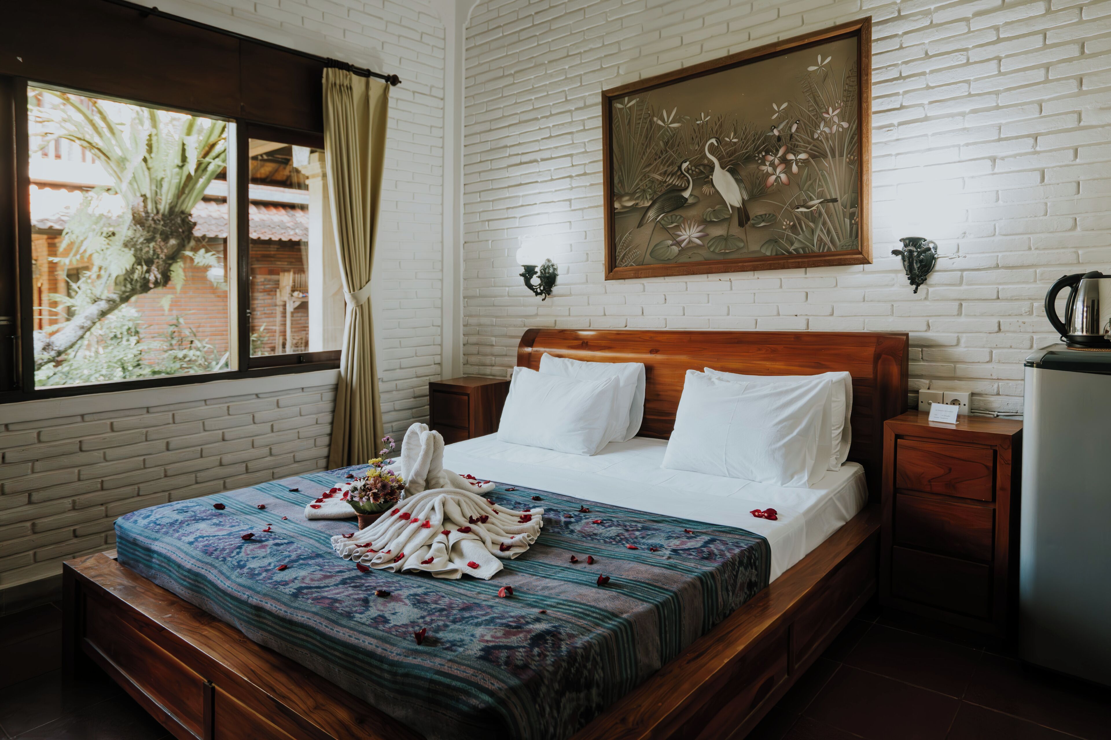 A hotel room with a brick wall, a wooden bed, a painting, a window with curtains, and a nightstand with a kettle.