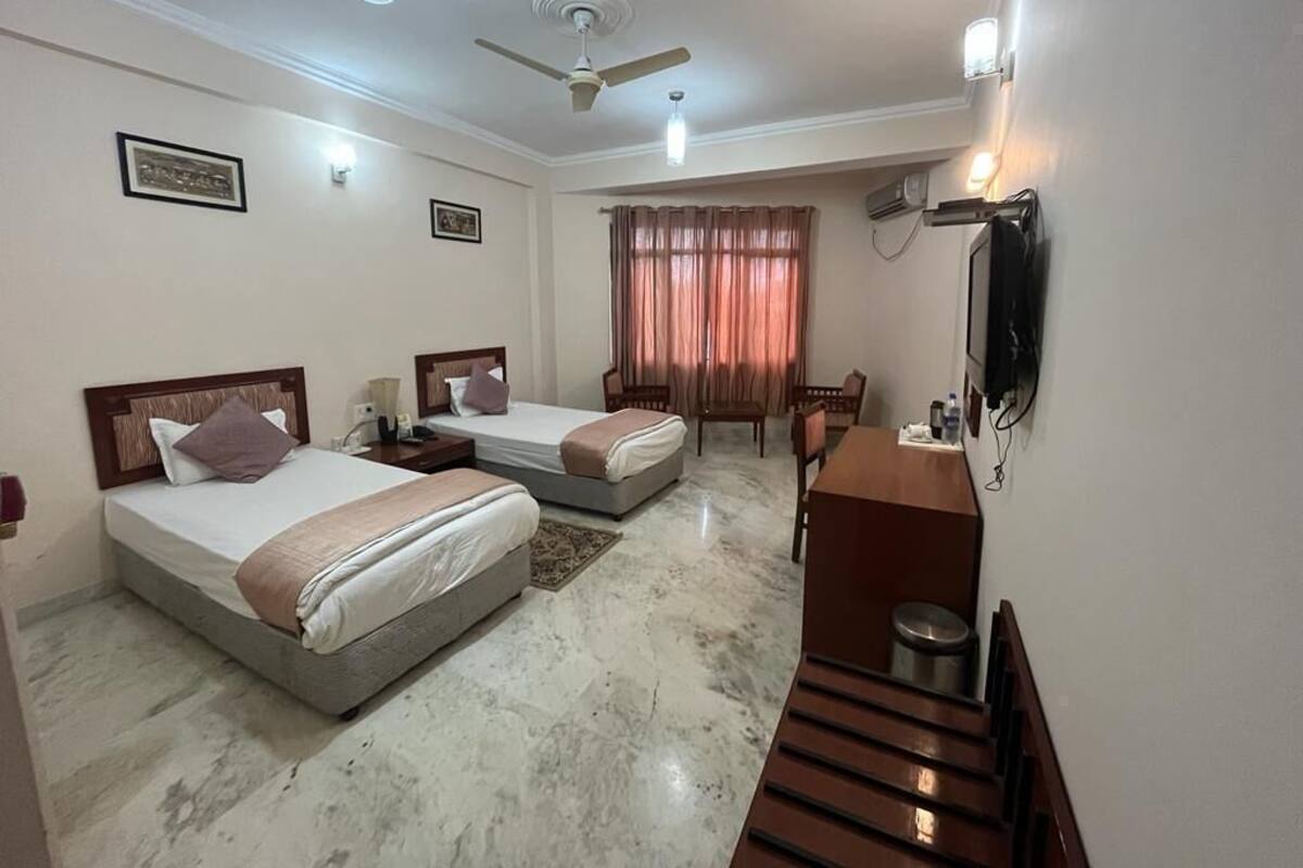 Quarto de hotel com duas camas, uma escrivaninha, uma cadeira, ventoinha no teto e televisão.