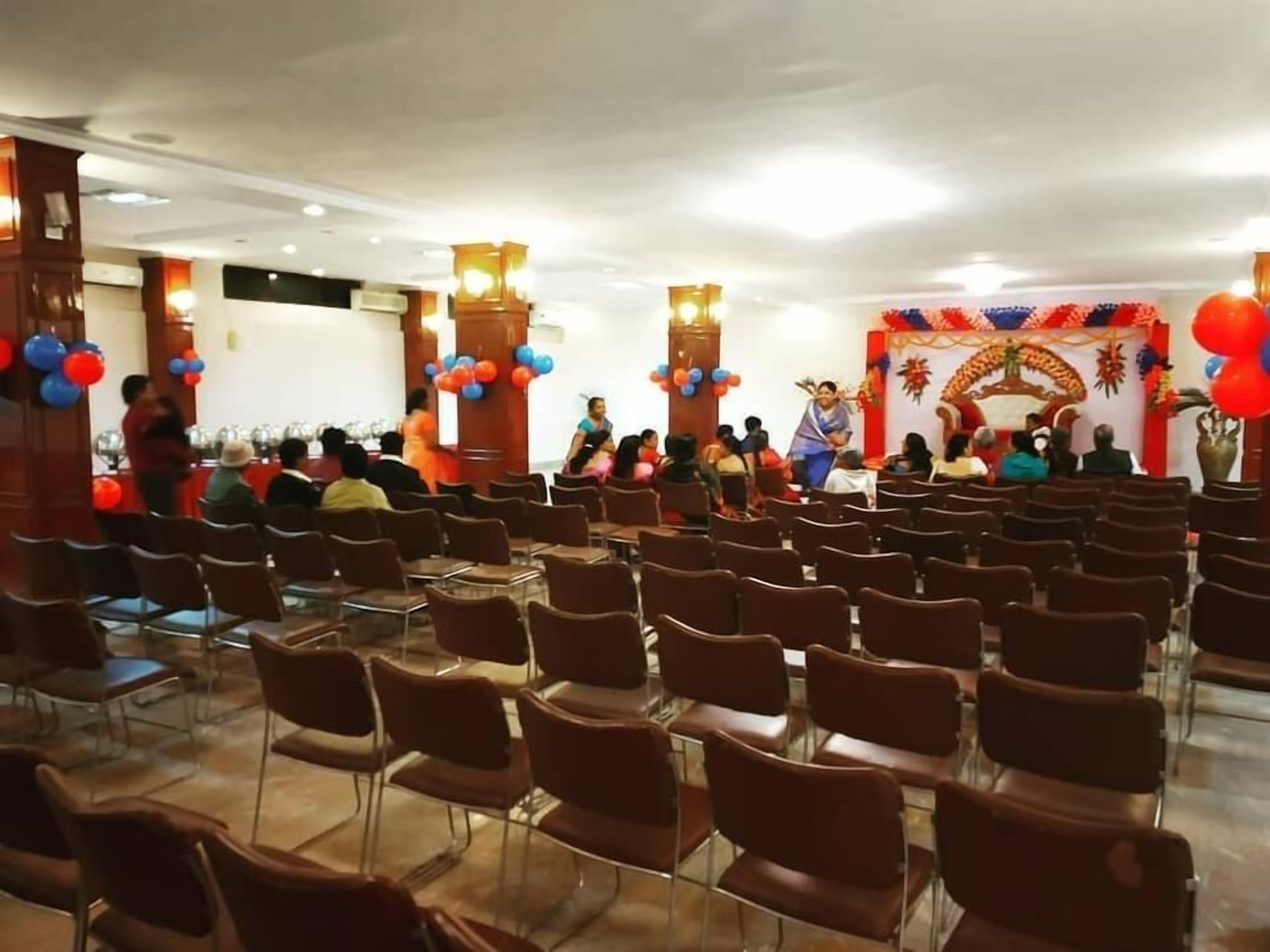Un salón de conferencias con filas de sillas, un escenario con un orador y un fondo decorativo.