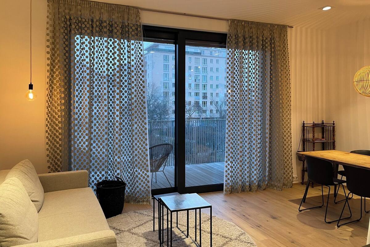 A modern living room with a sofa, a small table, a dining table with chairs, and a view through large glass doors.