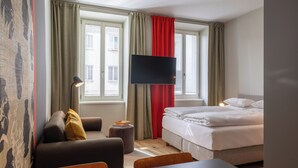 Habitación de hotel con cama, sofá, escritorio, televisor y ventana con cortinas.