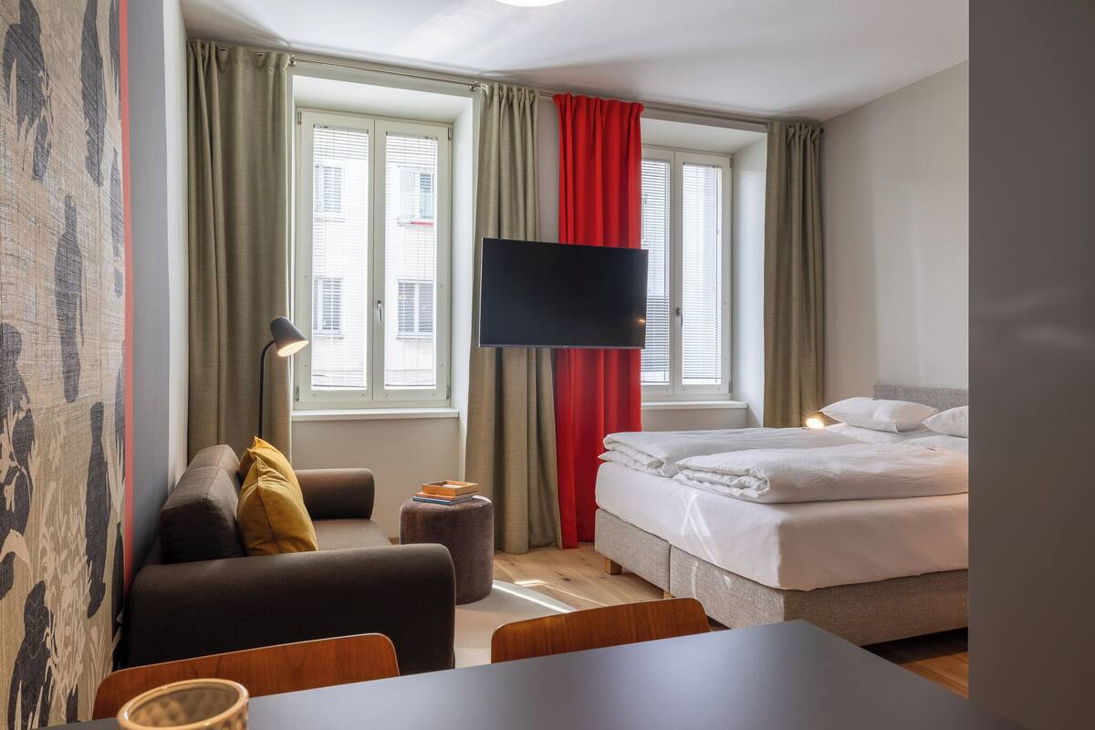 A hotel room with a bed, a sofa, a desk, a TV, and a window with curtains.