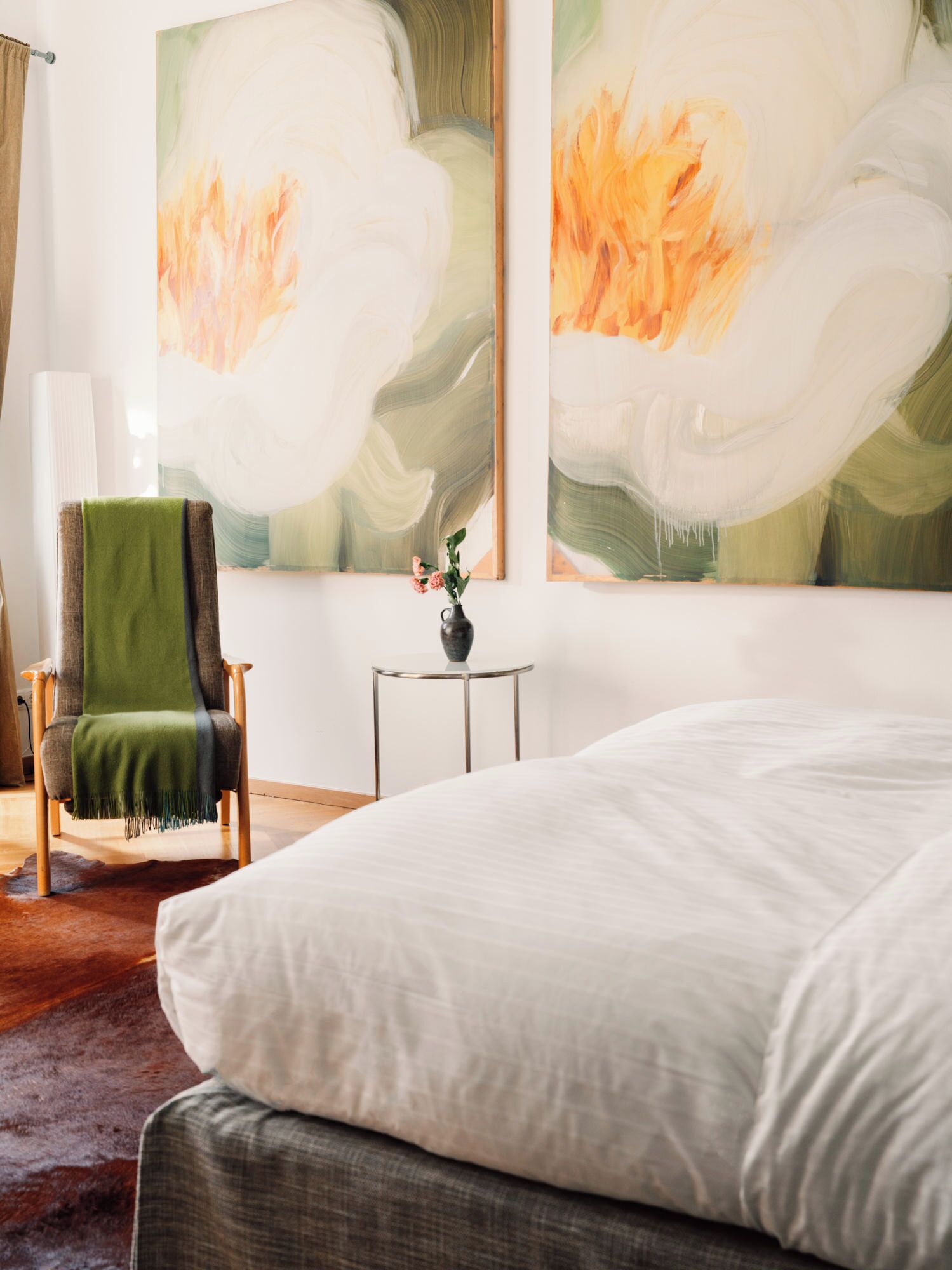 Ein Schlafzimmer mit einem Bett, einem Sessel, einem kleinen Tisch mit einer Vase sowie zwei großen abstrakten Bildern an der Wand.