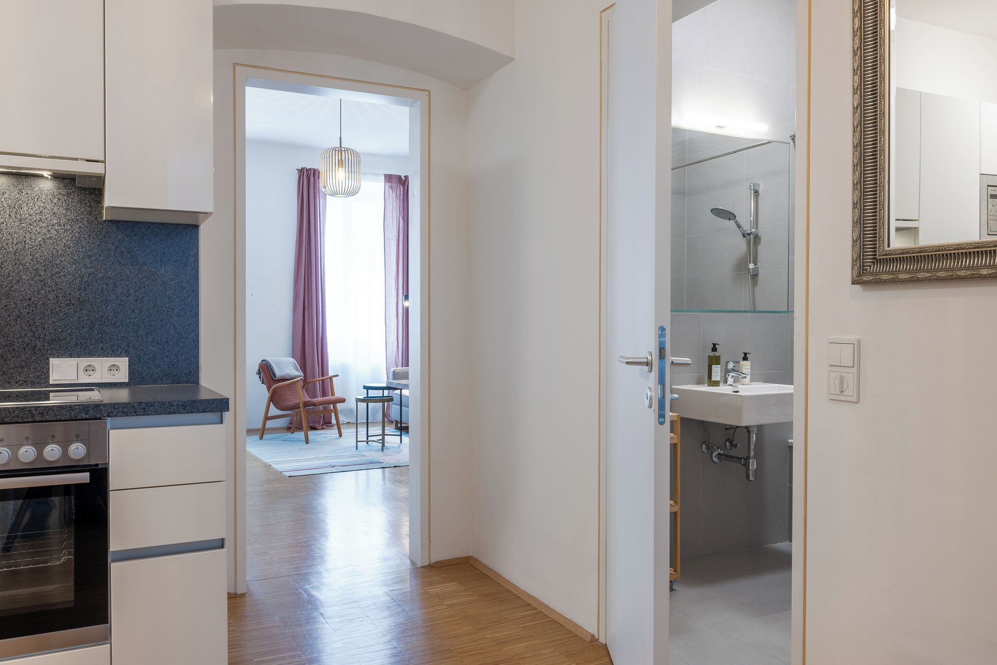 A modern kitchen with a built-in oven, a dining area with a chair and table, and a bathroom with a glass shower and sink.