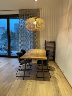 Un espace repas avec une table en bois, des chaises noires, un luminaire suspendu et une vue dégagée sur l’extérieur grâce à une grande fenêtre.