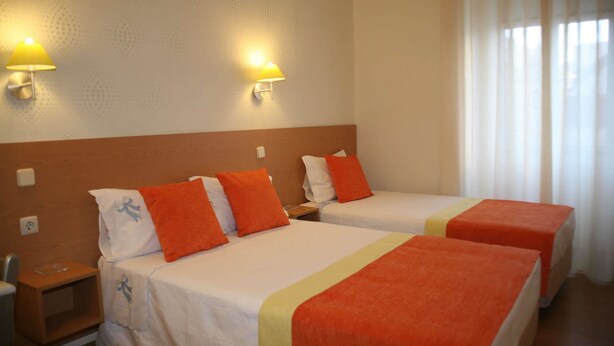 Een hotelkamer met twee bedden, elk voorzien van oranje kussens en een witte sprei. Aan weerszijden van de bedden hangen twee wandlampen.