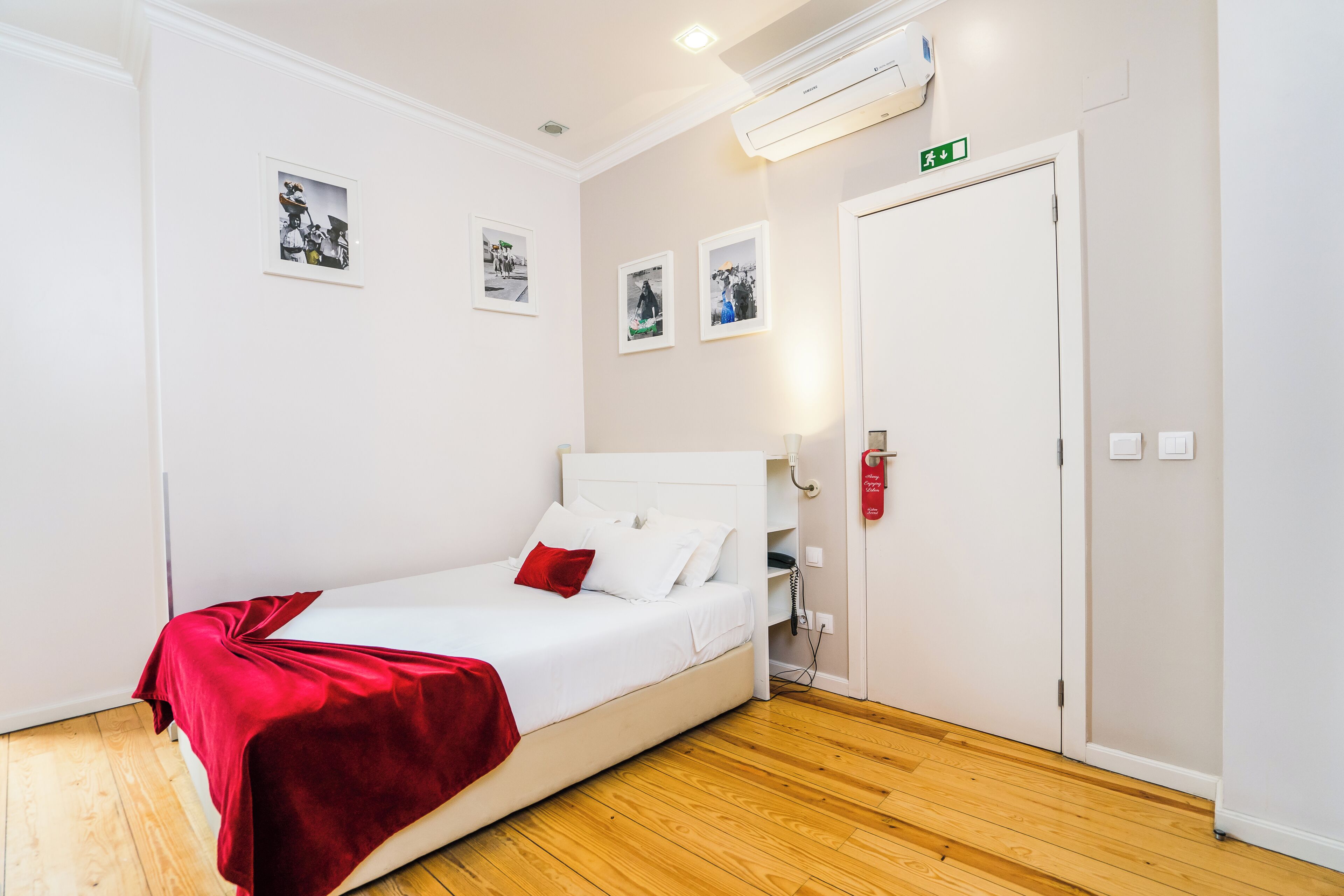 A hotel room with a white bed, red throw blanket, wooden flooring, and three framed pictures on the wall.