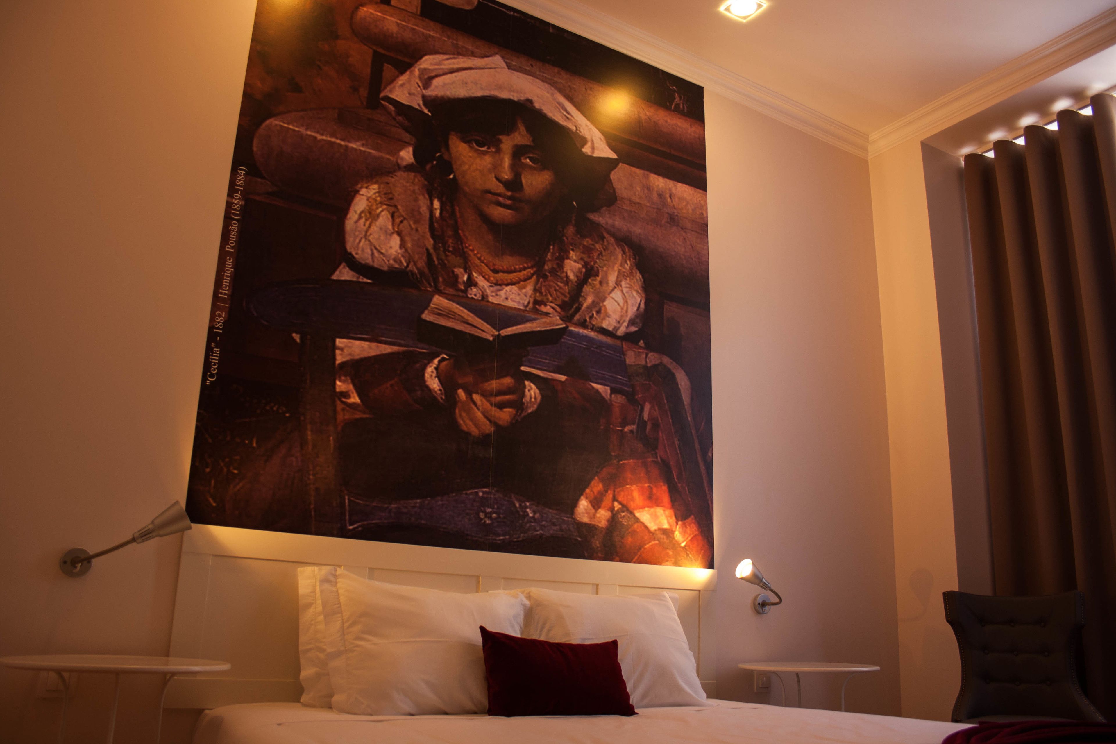 A hotel room with a large painting on the wall, a bed with white bedding and a red pillow, a small bedside table, and a chair.