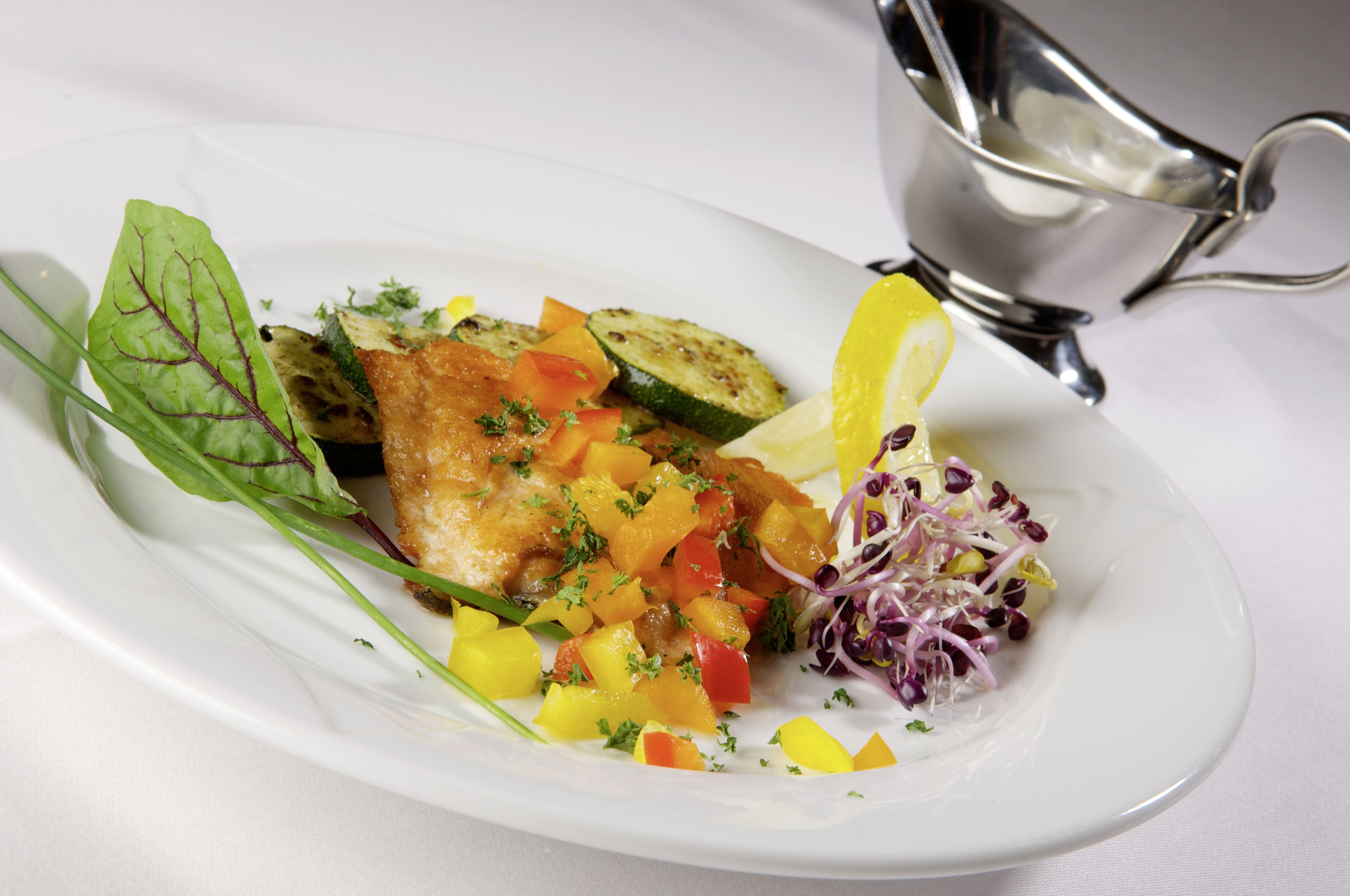 Un plato blanco con un filete de pescado, mango en cubos, pepino y una ensalada, acompañado de una salsera plateada.