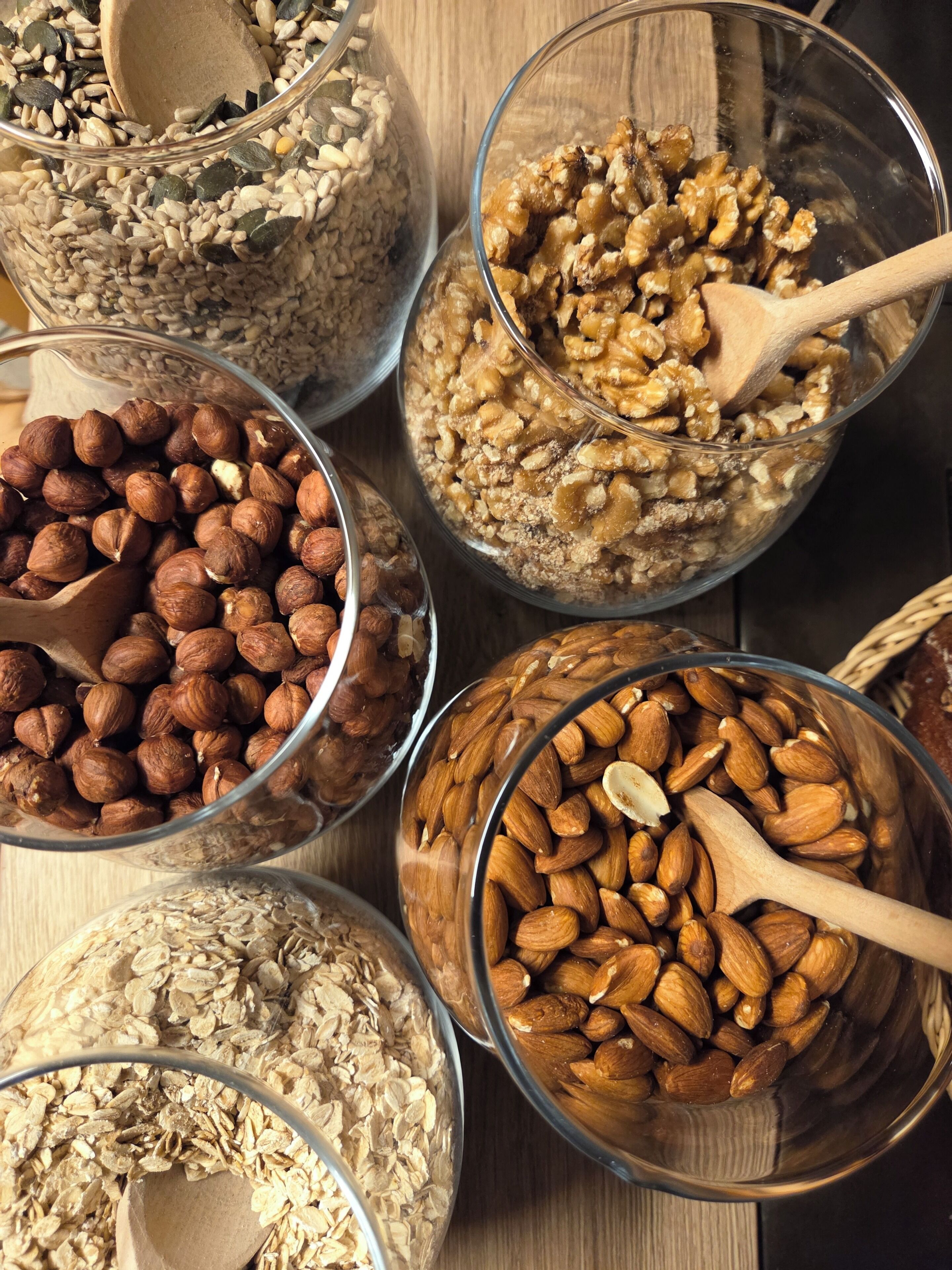 Various bowls of nuts and grains, including almonds, walnuts, and oats, with wooden spoons in them.
