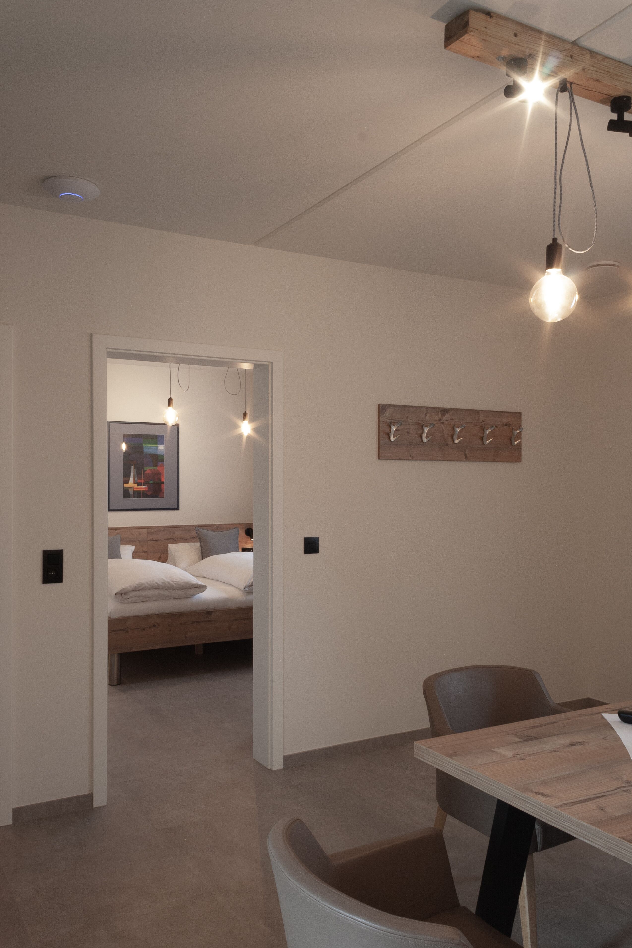 A modern bedroom with a wooden headboard, a bed with white bedding, a dining table with chairs, and a wall-mounted light fixture.