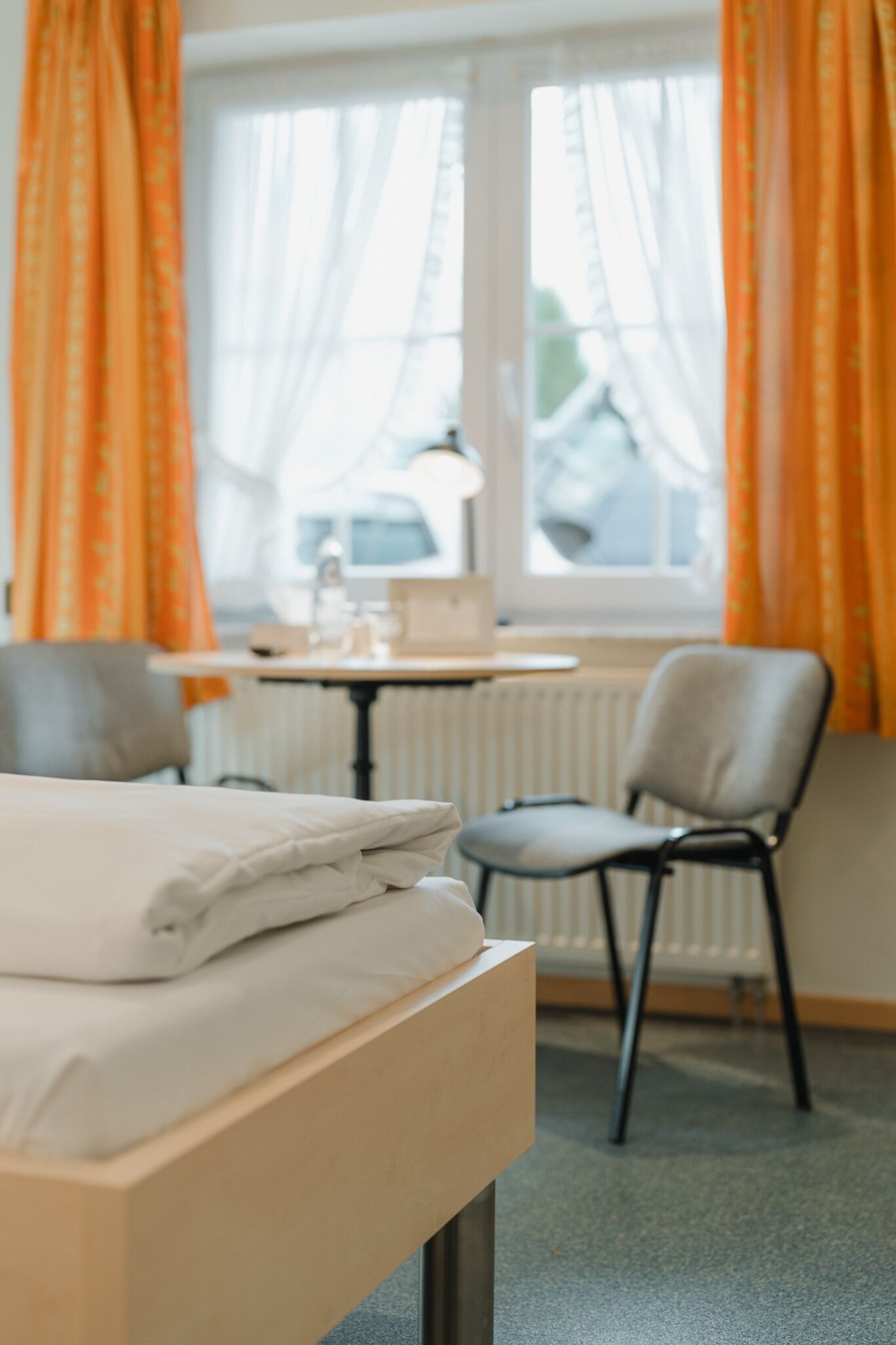 A hotel room with a bed, a chair, a small table, and a window with curtains.