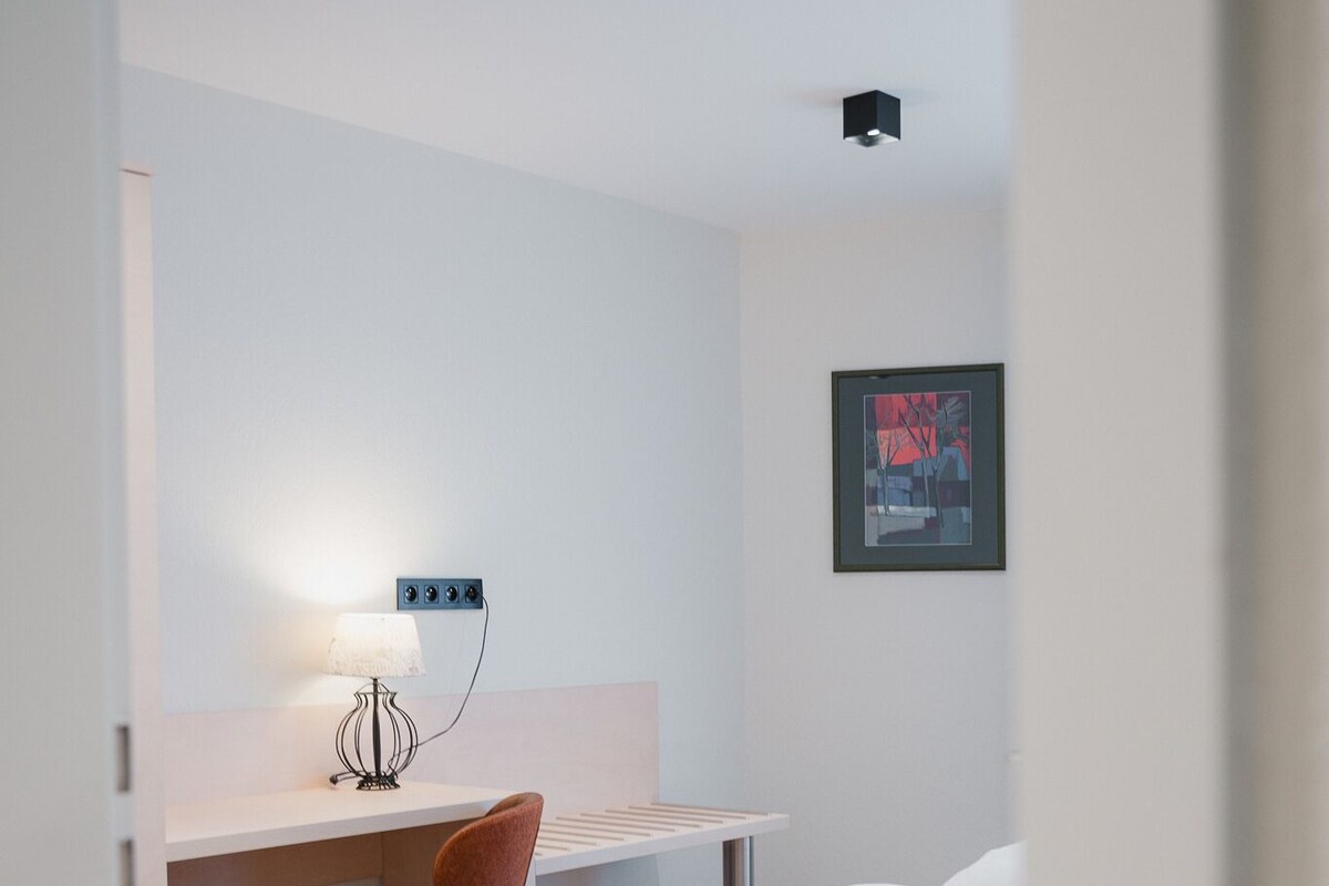 Habitación de hotel con suelo de madera, cama con ropa de cama blanca, escritorio con silla y una lámpara fijada a la pared.