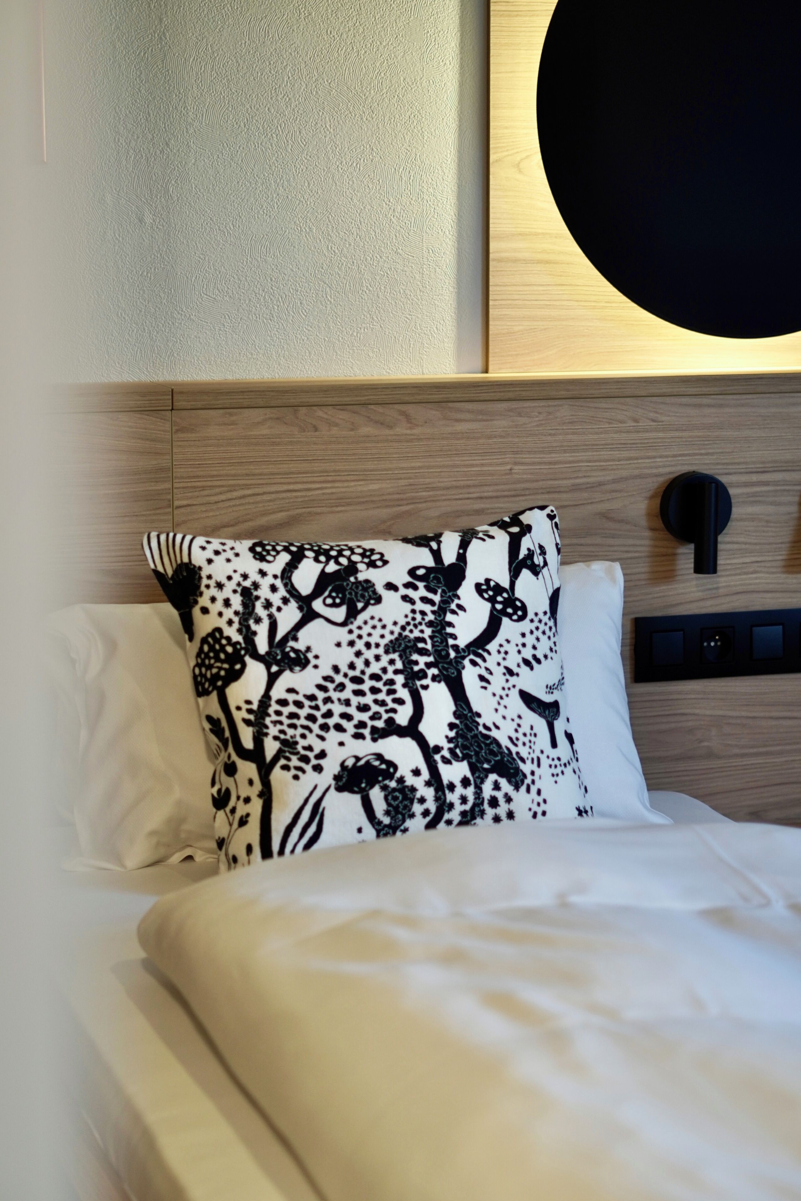 Una cama bien hecha con ropa de cama blanca y una almohada floral negra, un cabecero de madera y una lámpara de pared.