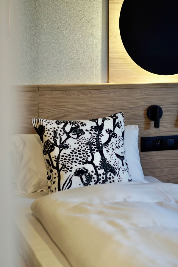 Una cama bien hecha con ropa de cama blanca y una almohada floral negra, un cabecero de madera y una lámpara de pared.