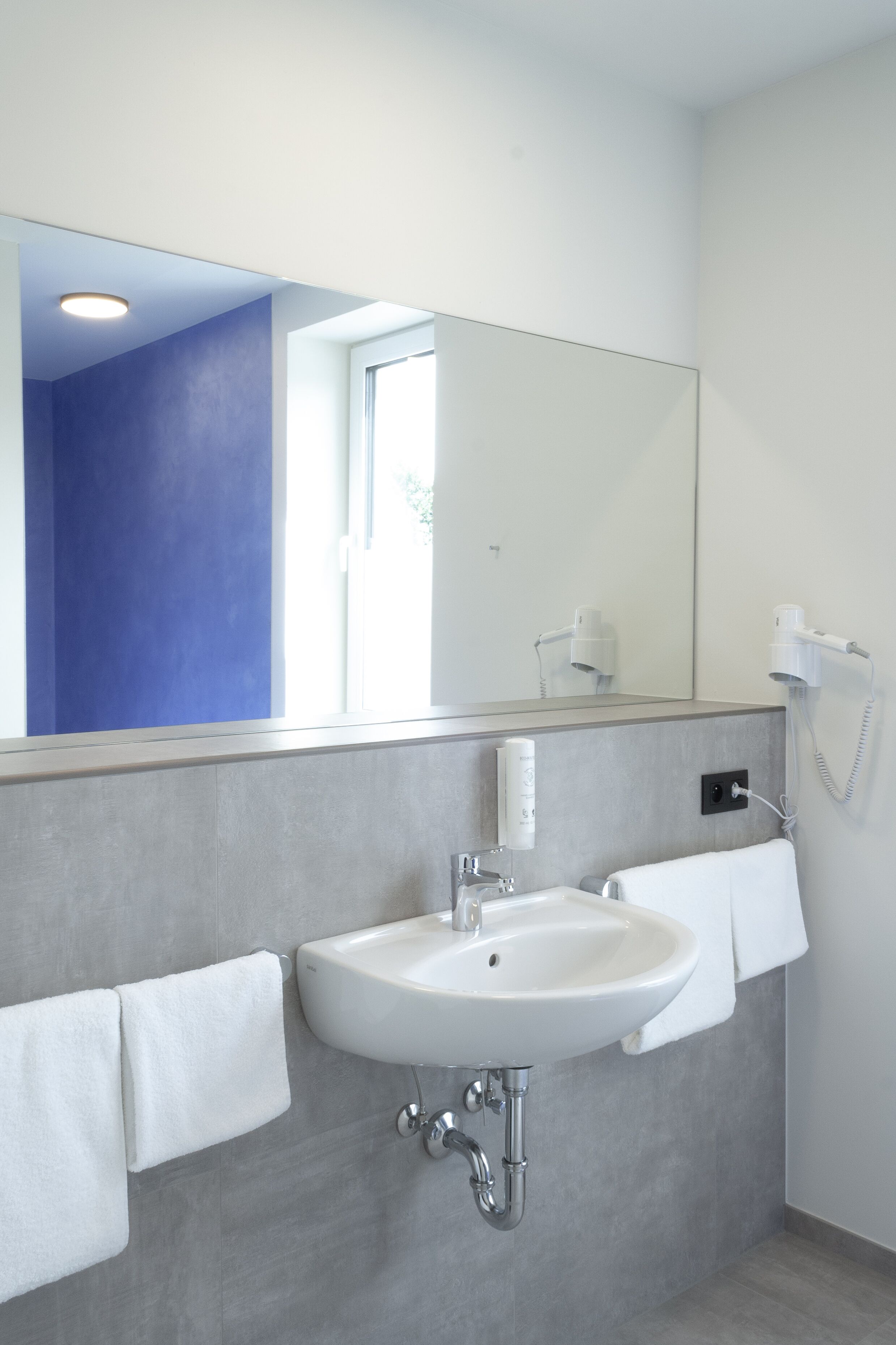 Un baño con lavamanos blanco, un espejo y un secador de pelo en la pared.