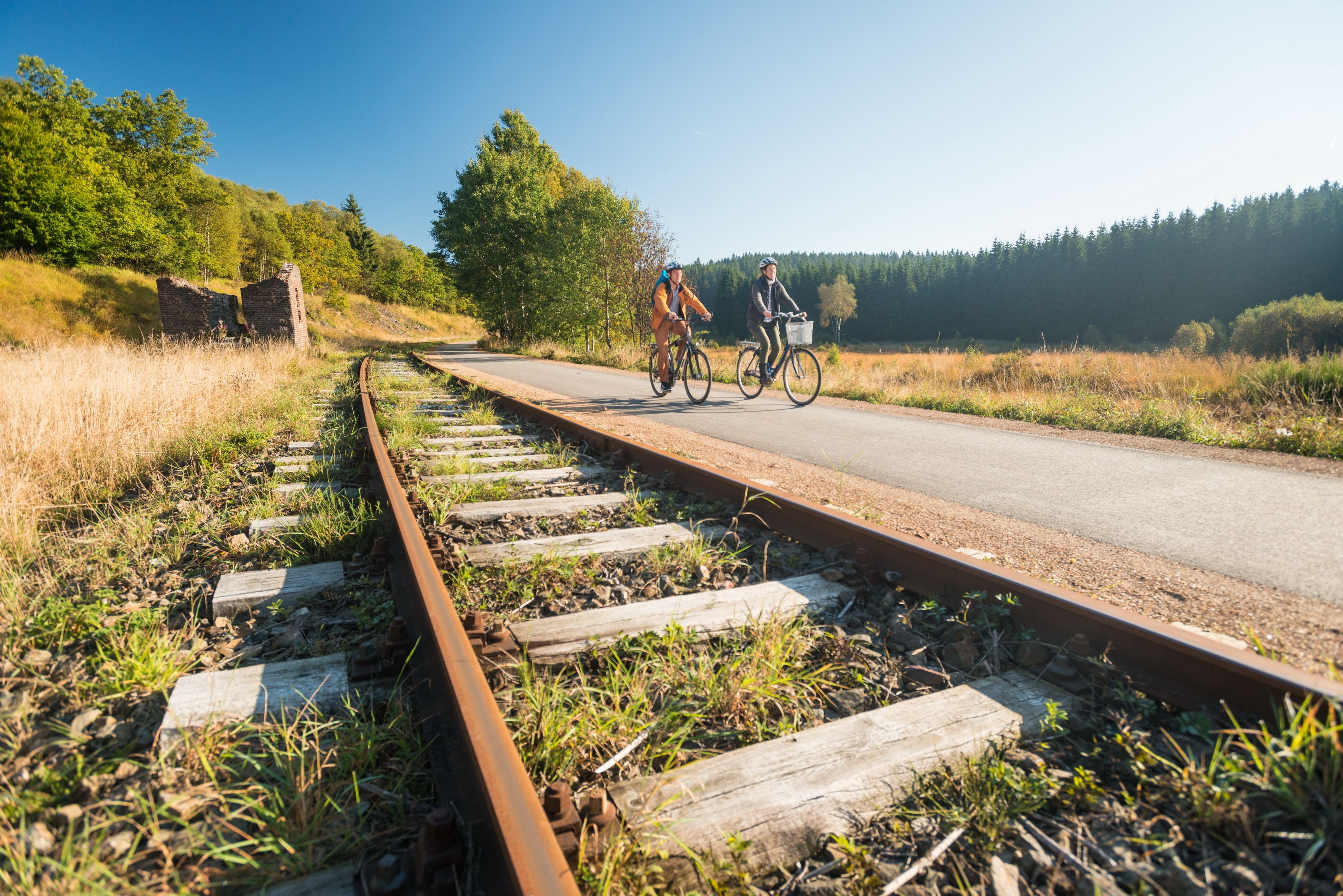 Una vía de tren con durmientes de madera y rieles de metal, un camino pavimentado a su lado, y dos ciclistas pedaleando por el camino.