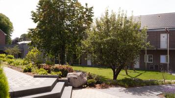Stroll through the beautifully landscaped grounds, surrounded by lush plants and a majestic tree, leading to a modern building with a brick façade and large windows.