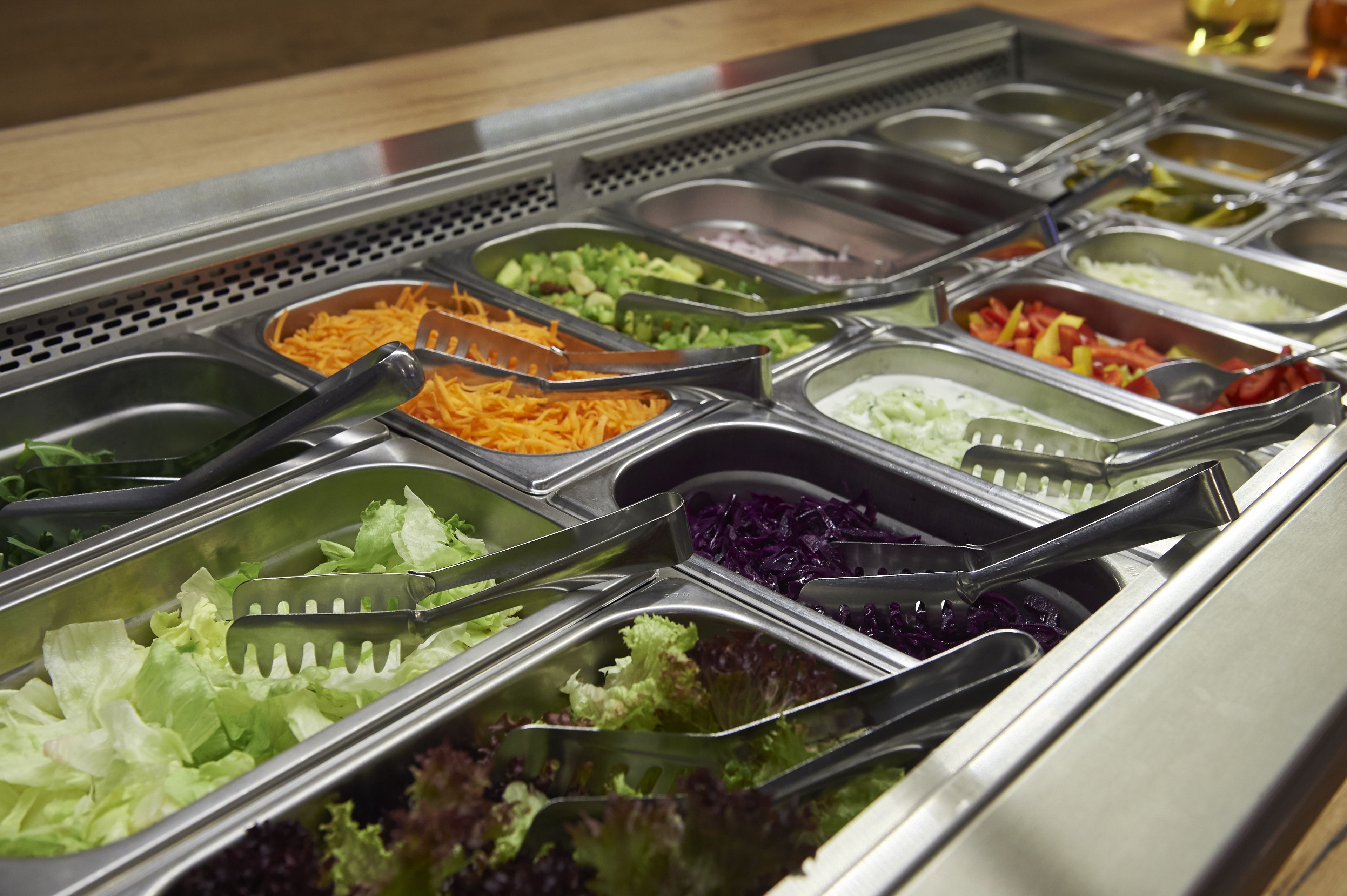 Ein Buffet mit verschiedenen Speisen in separaten Behältern, darunter geriebener Käse, gemischtes Gemüse und verschiedene Salatsorten.
